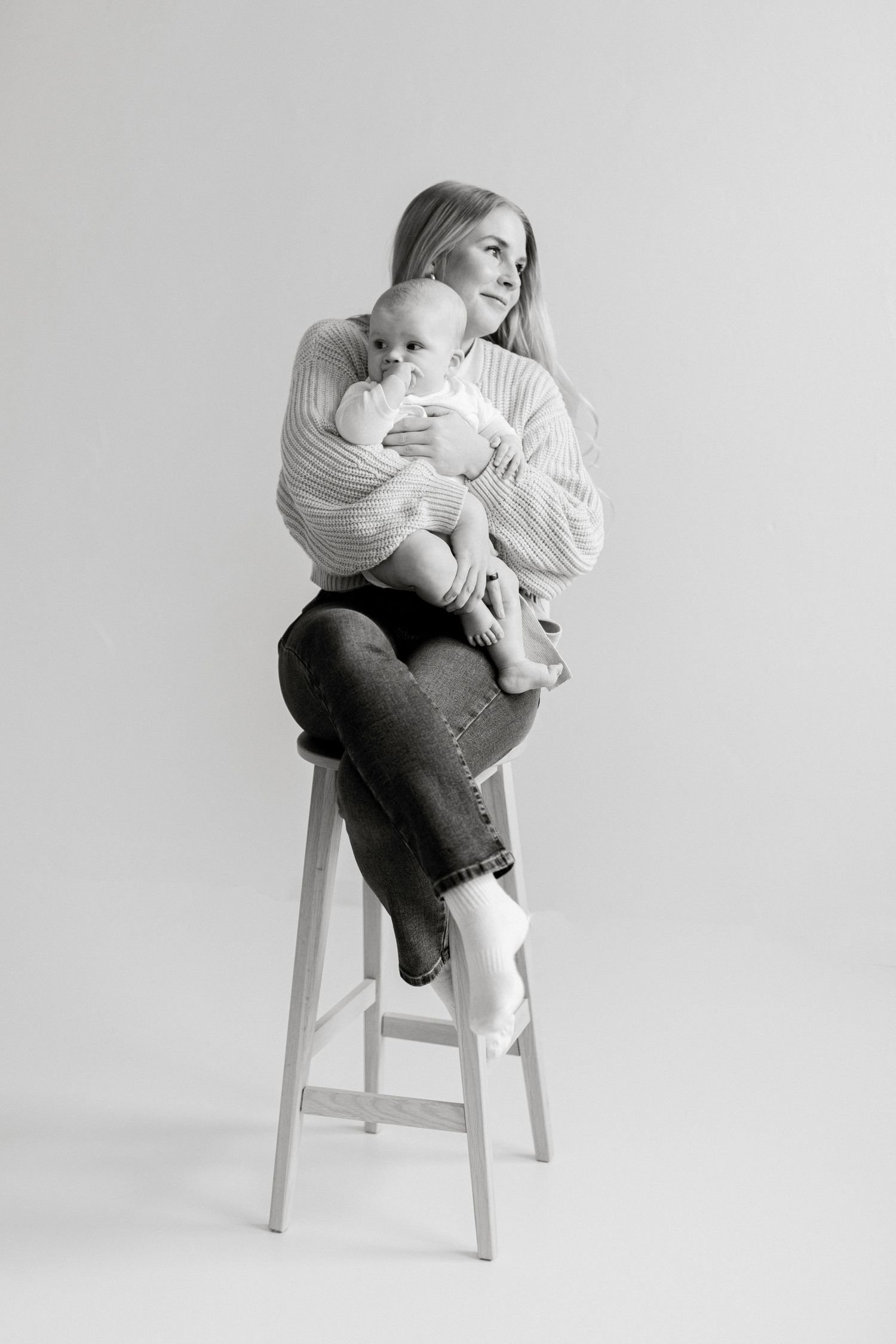 Black and white studio photo of a young mother sitting on a stool, tenderly holding her newborn baby.