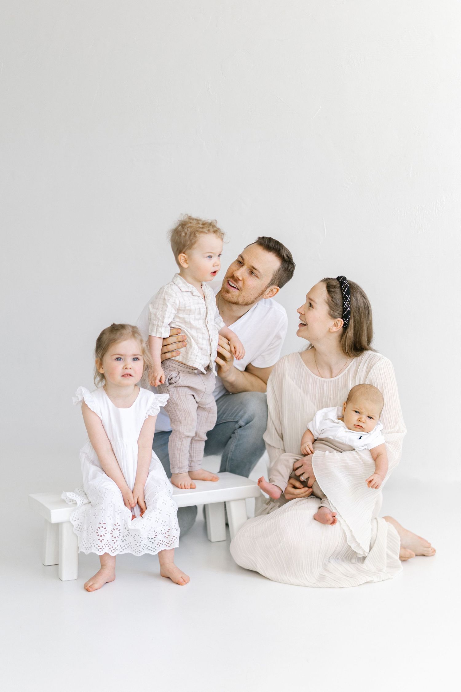Happy family of five with newborn baby posing together in white outfits against a light background.