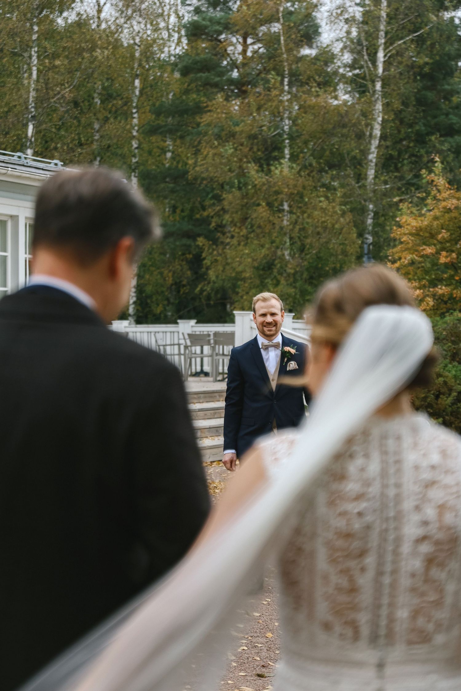 Smiling groom in navy suit watches bride and guest approach during outdoor autumn wedding ceremony.