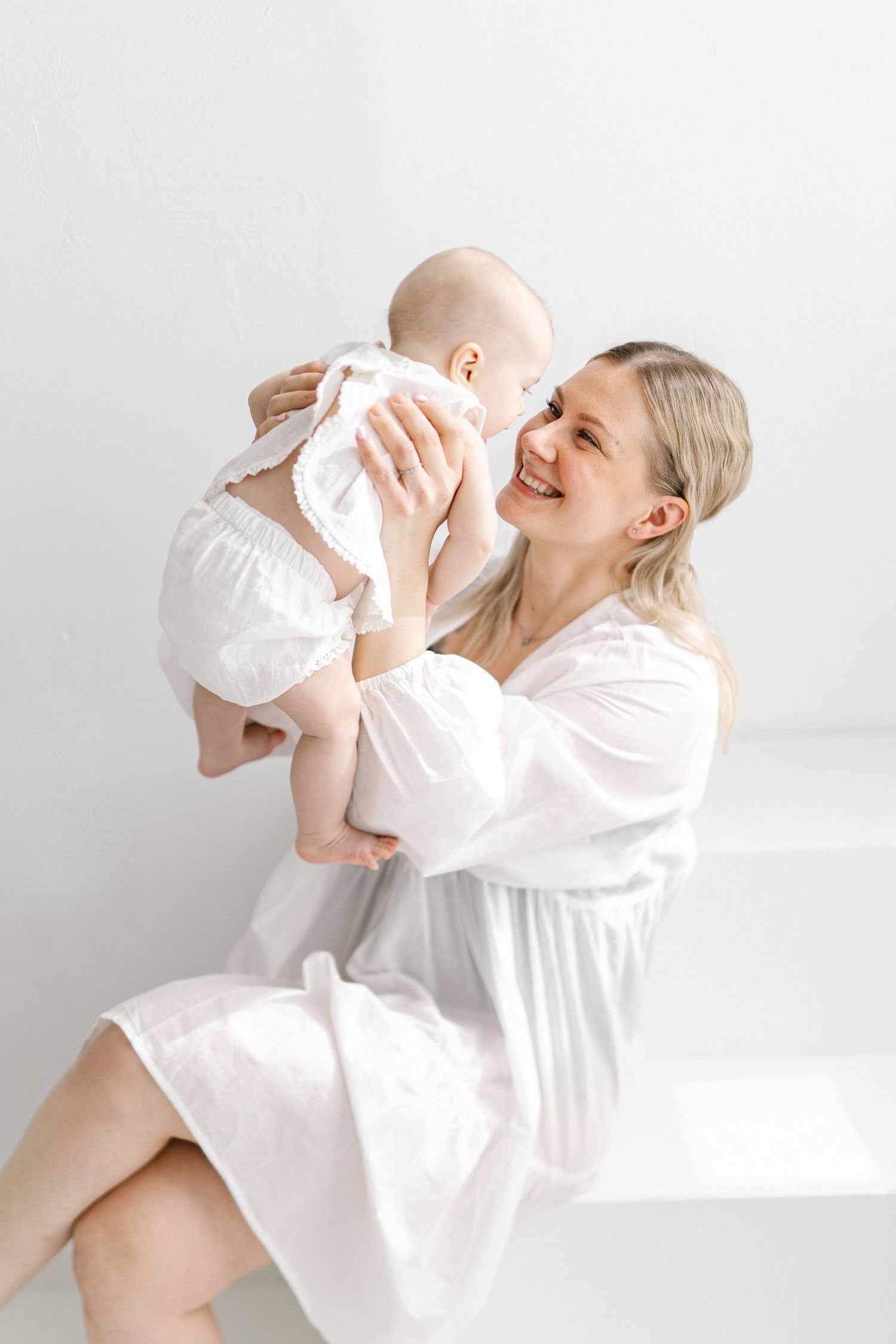 Smiling blonde mother in white dress holding baby in white outfit against bright white background.