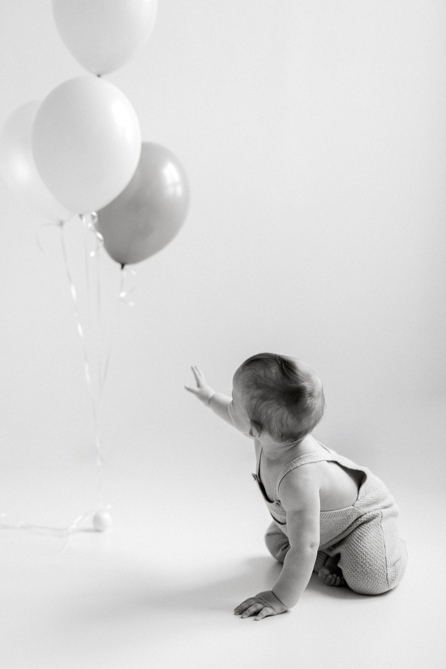 Black and white photo of a baby in overalls reaching toward three balloons while sitting on a white studio floor.