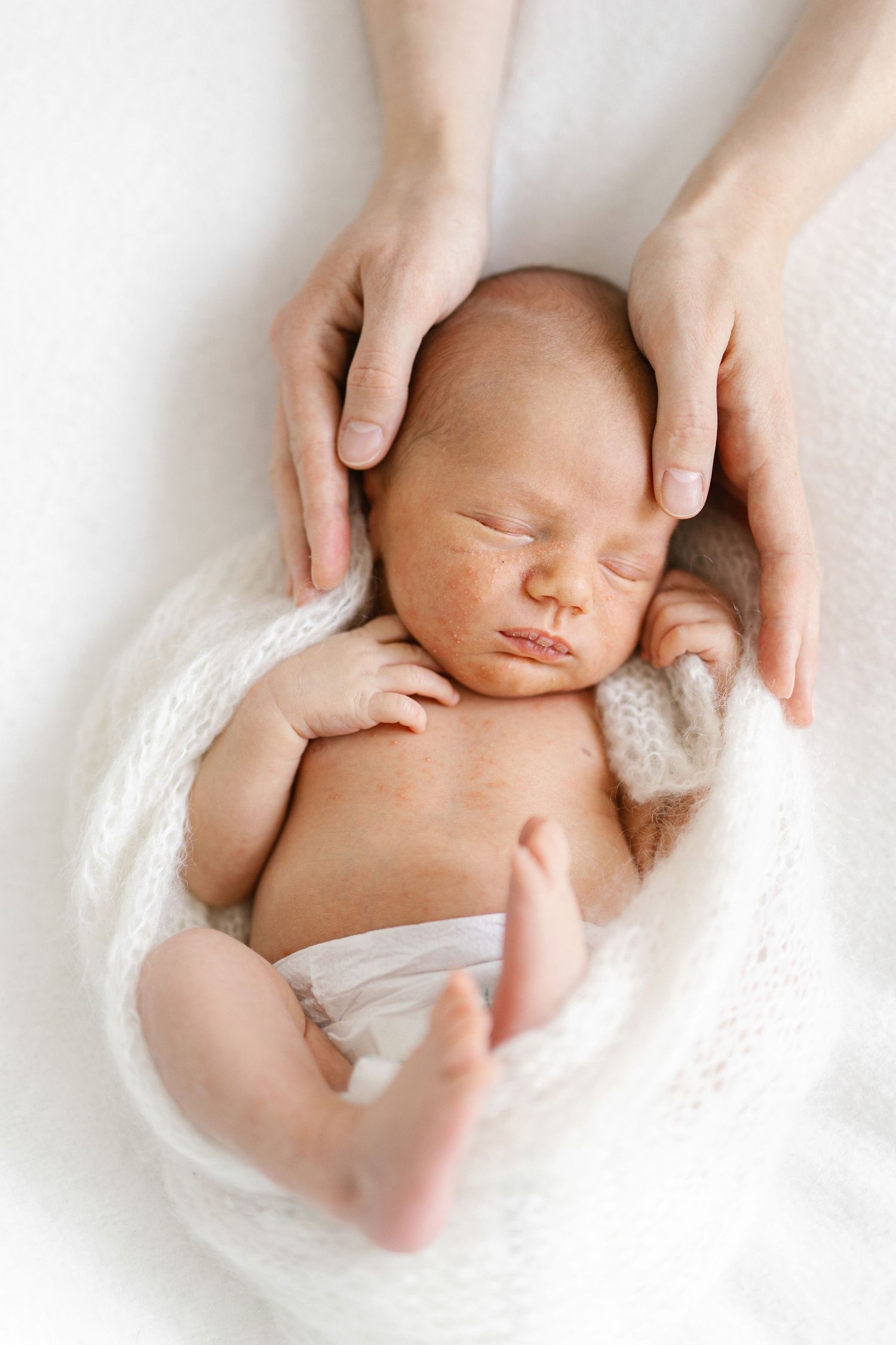 Newborn baby sleeping peacefully wrapped in white knit blanket while mother's hands gently cradle the infant's head.