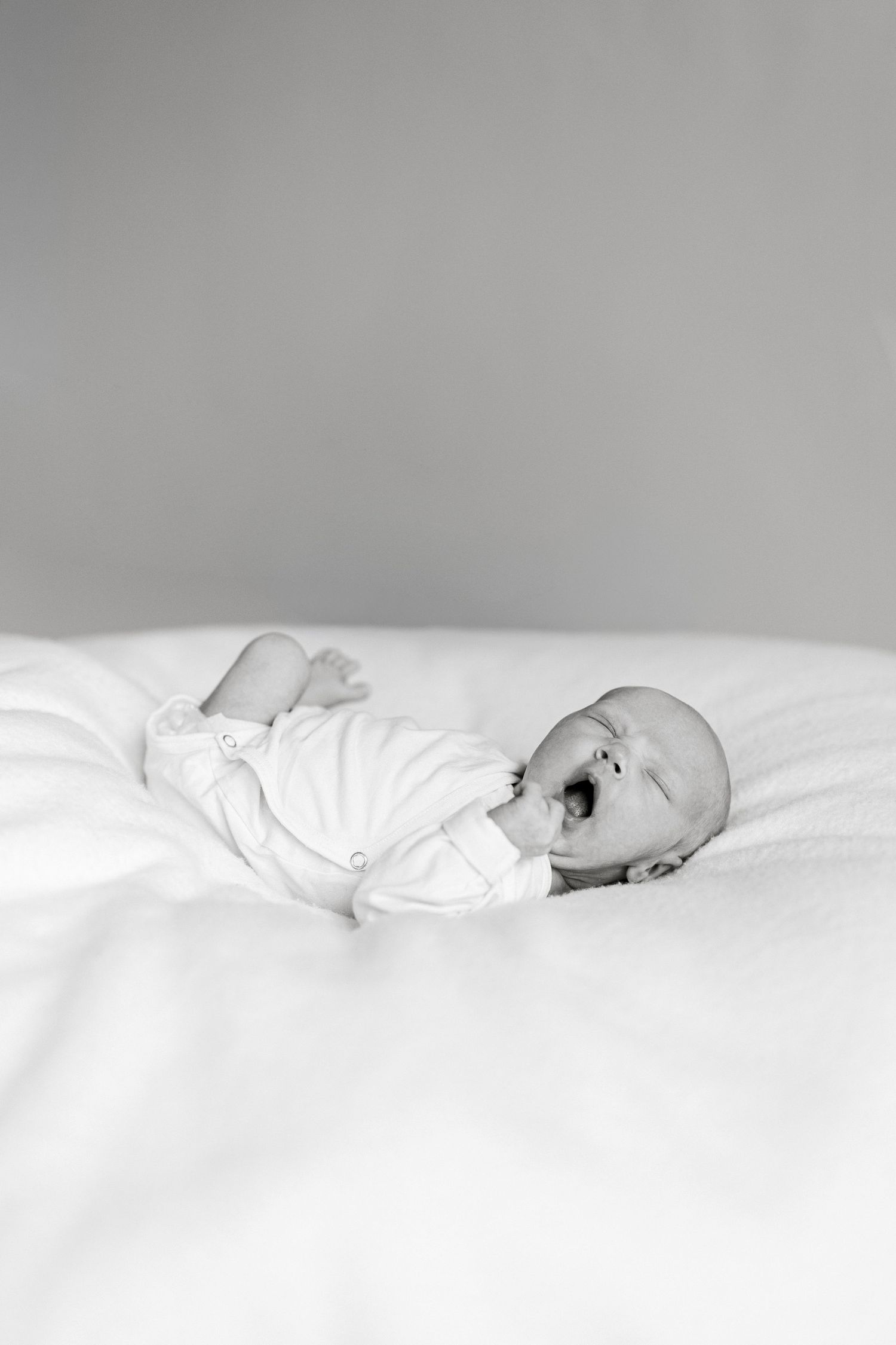 Black and white photo of a newborn baby yawning while lying on a white blanket in a onesie.
