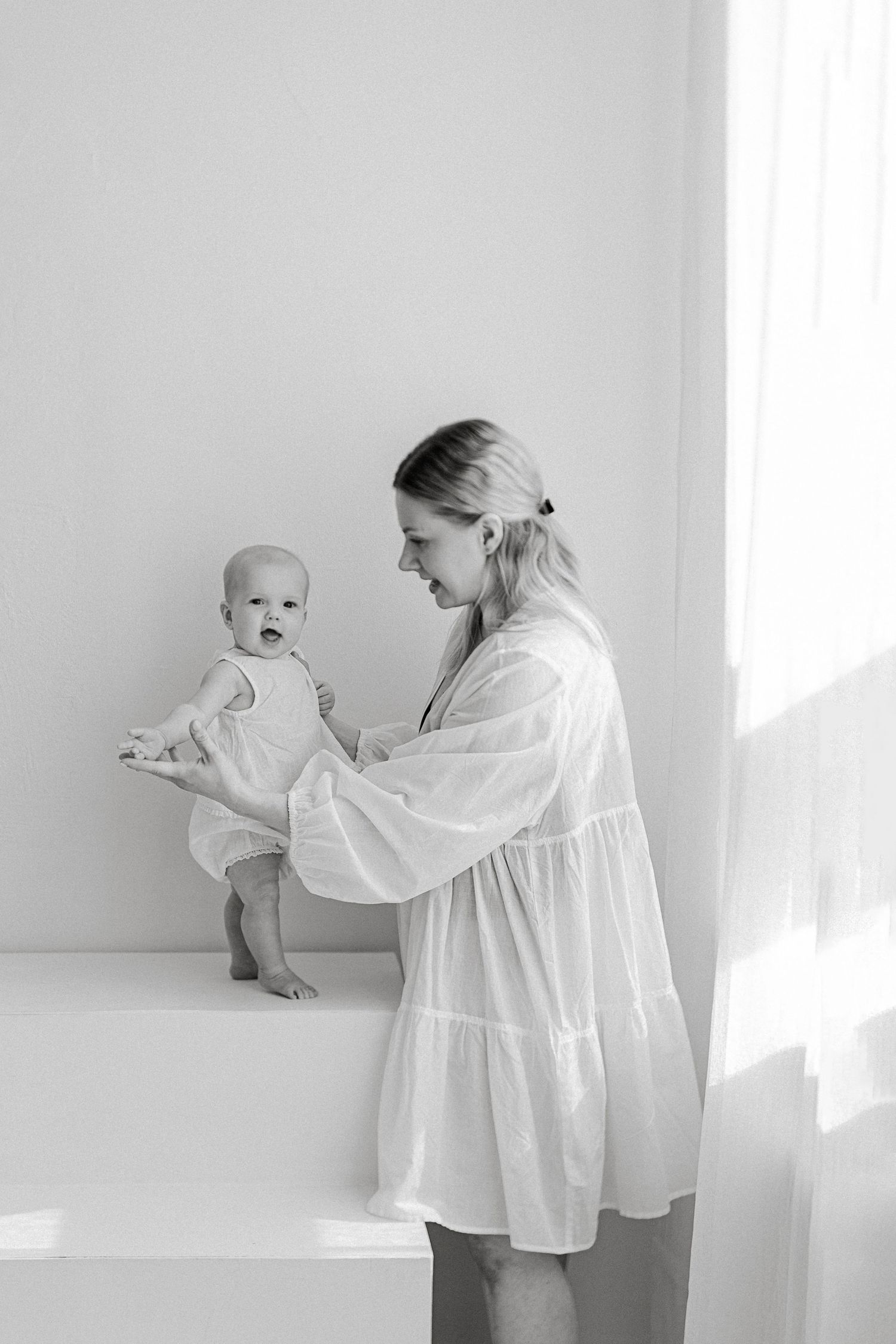 Black and white photo of a mother in white dress holding her baby on a white surface near a bright window.