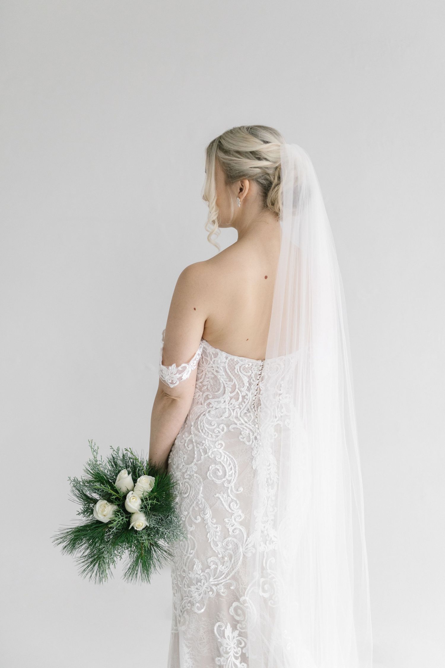 Back view of bride in detailed lace dress holding white bouquet against neutral background.