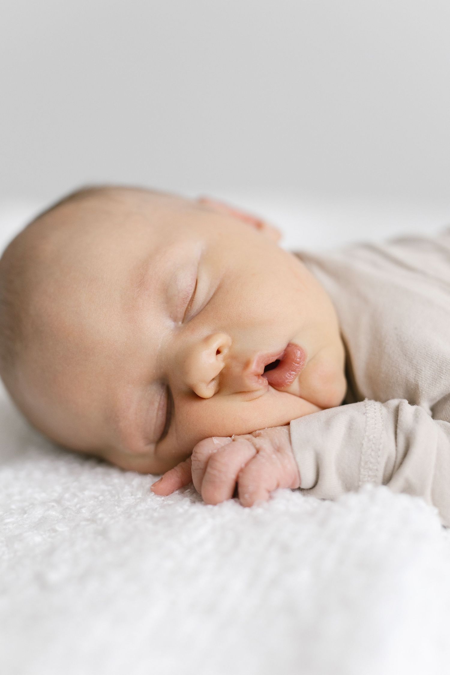 Side view of sleeping newborn in neutral clothing on white textured surface.
