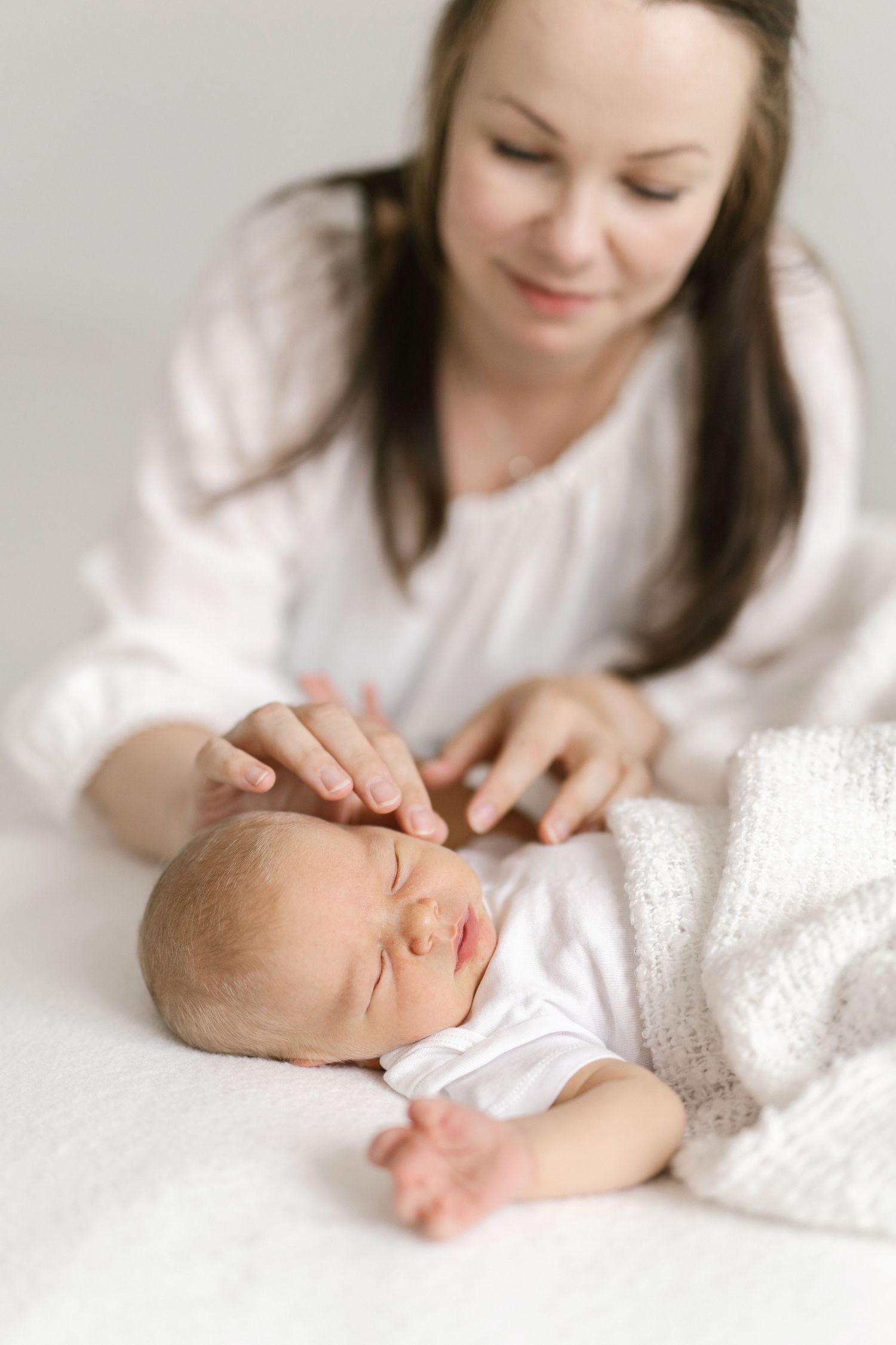 A mother tenderly strokes her sleeping newborn baby wrapped in a white knit blanket on a soft white surface.