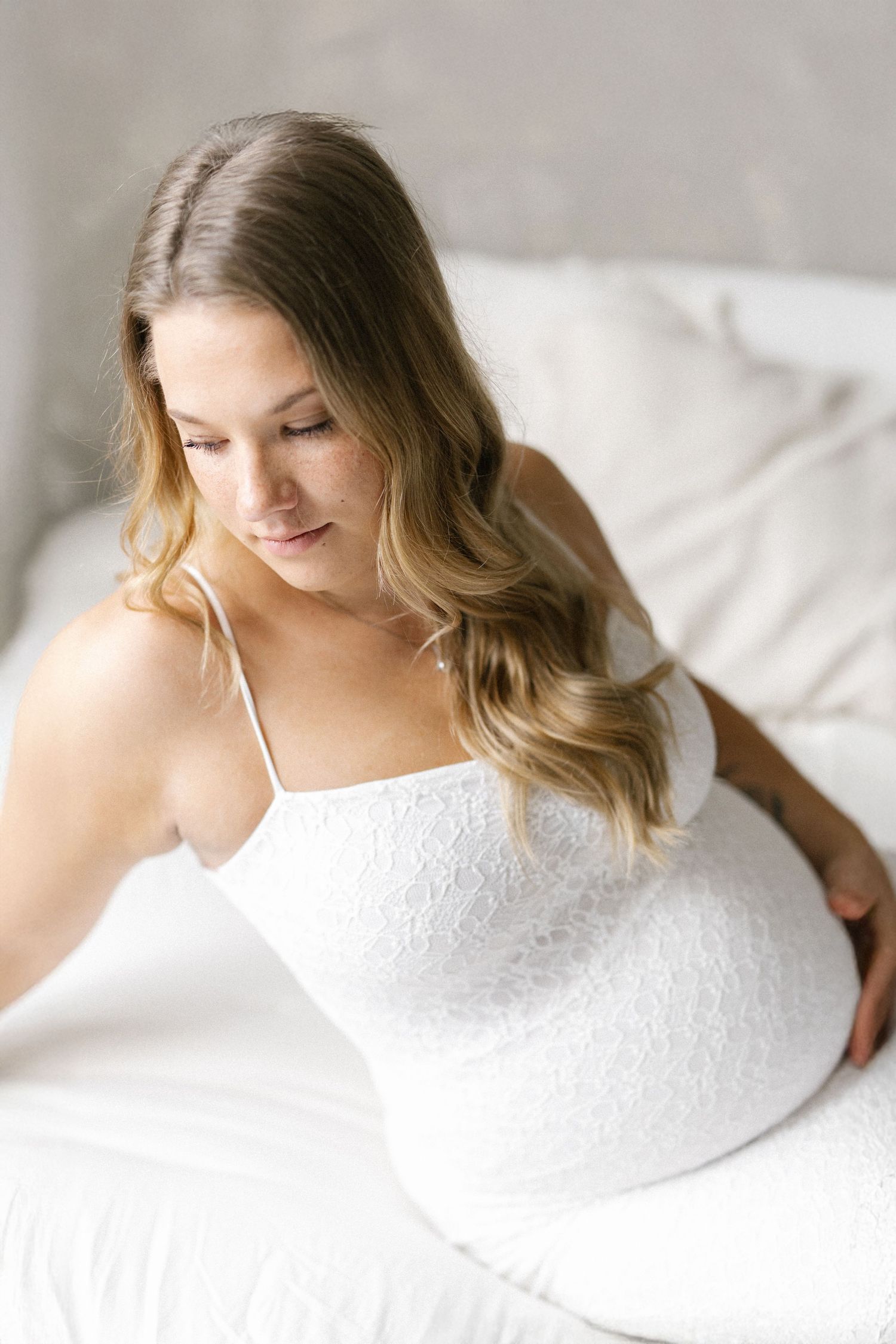 Pregnant woman in white lace dress sitting on bed, looking down at baby bump with long wavy hair.