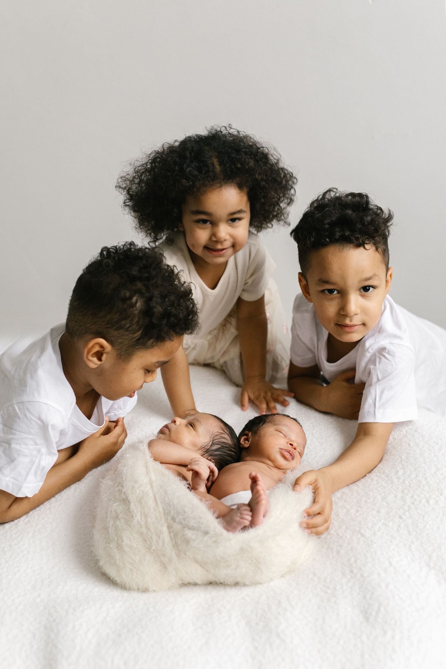 Three children in white outfits lovingly surround two swaddled newborn twins during a studio portrait session.