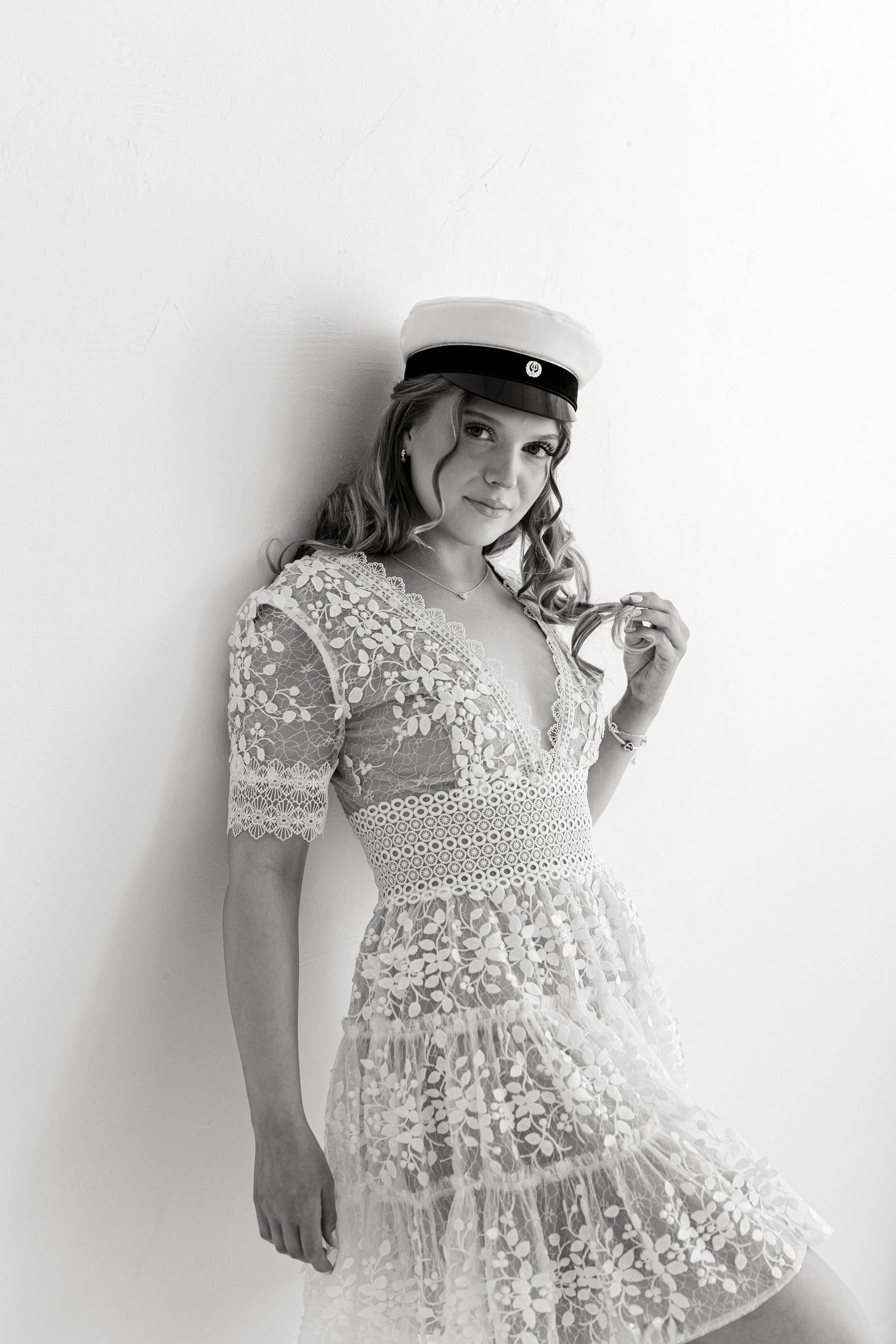 Black and white photo of a young woman in lace dress and sailor cap leaning against a white wall.