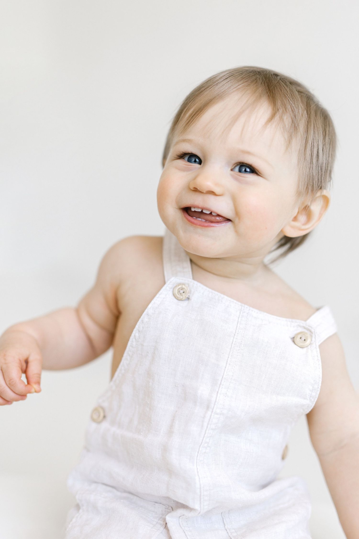 Smiling baby with blue eyes wearing white linen overalls against a light background.