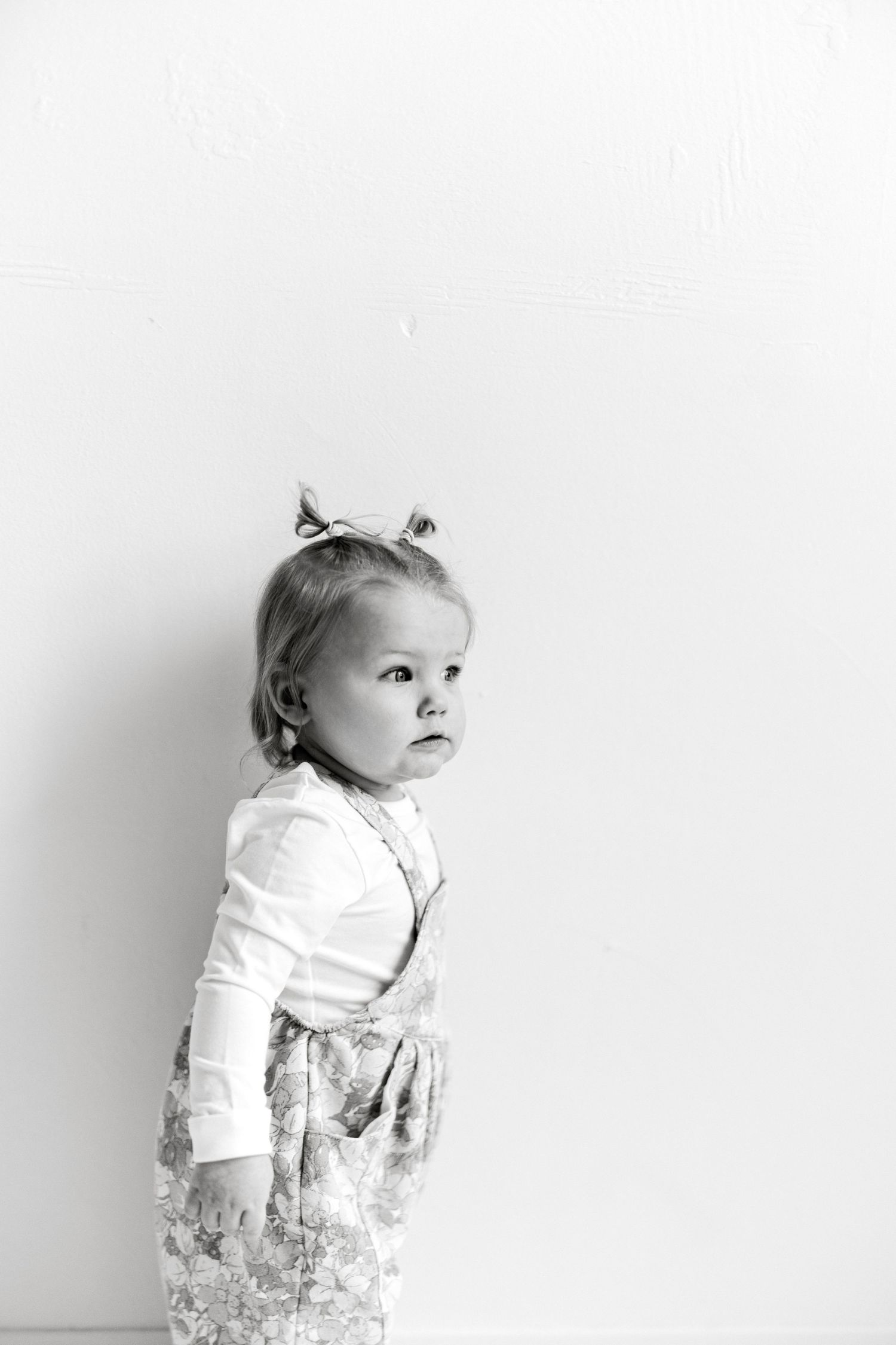 Black and white photo of a toddler girl in a floral dress with a bow in her hair, looking up curiously.
