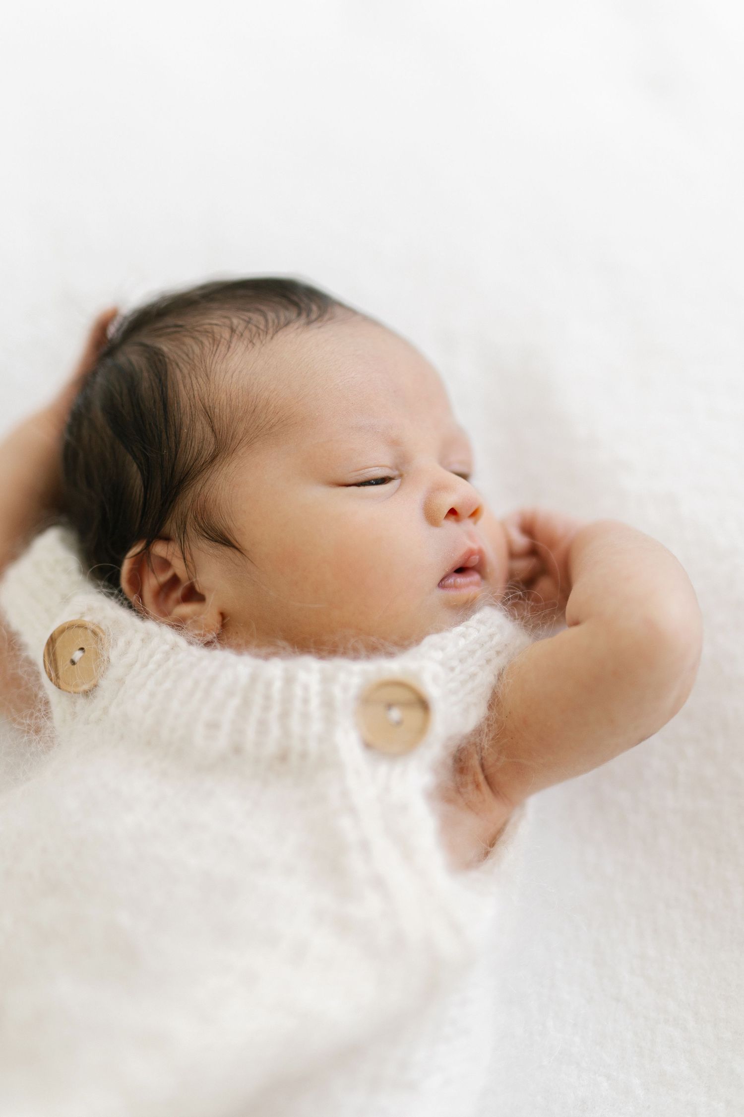Sleeping newborn baby in a white knit romper with wooden buttons, lying on a soft white blanket.