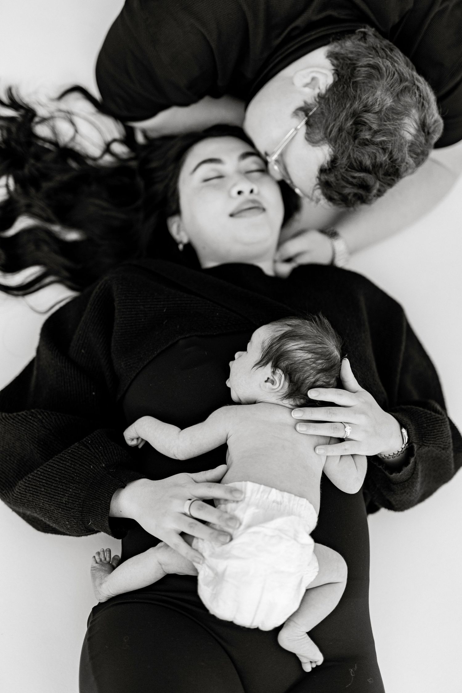 Black and white photo of parents lying down with newborn baby resting on their chests in a tender family moment.