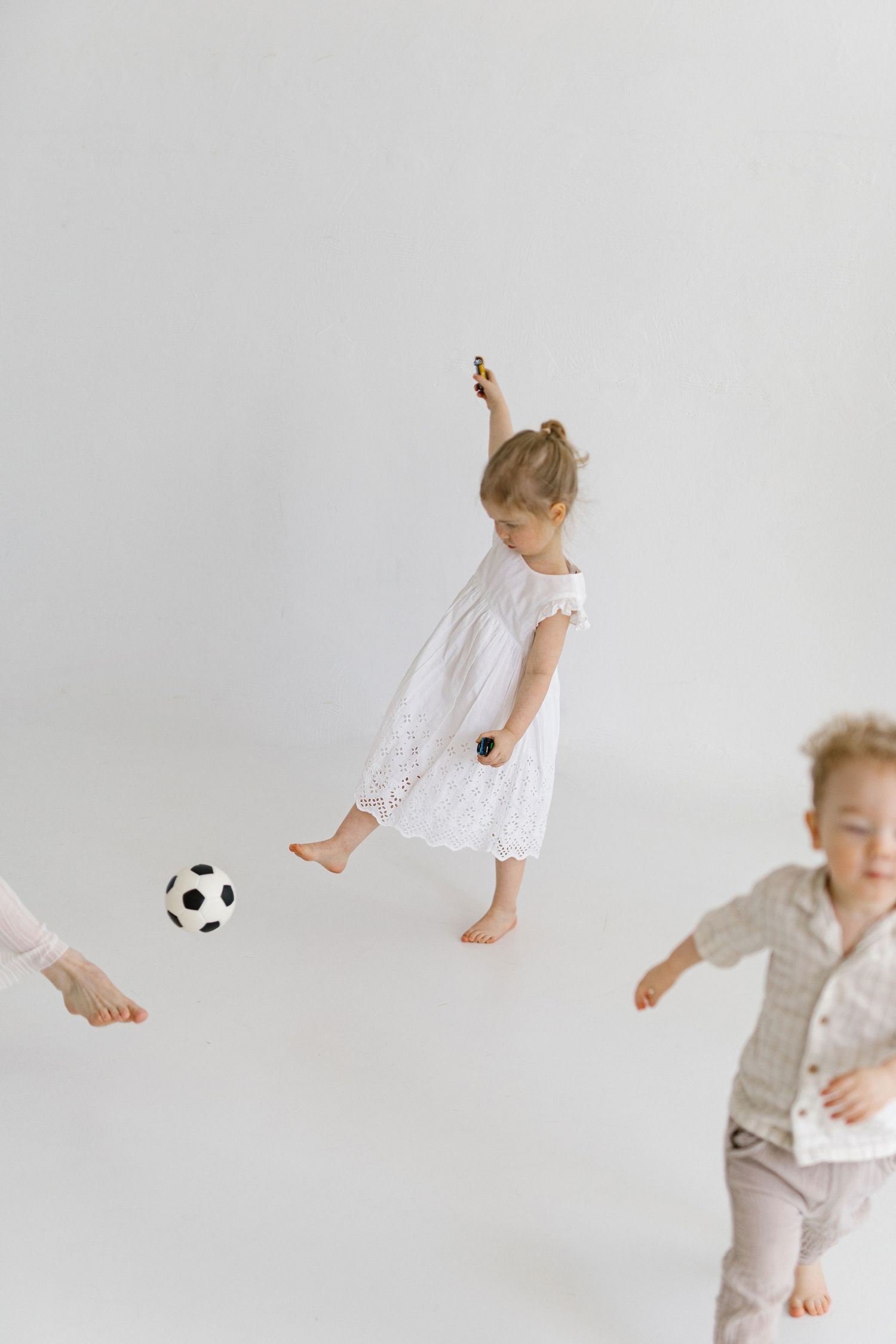 Two young children playing barefoot with a soccer ball in a bright white studio, girl in white dress kicking the ball.