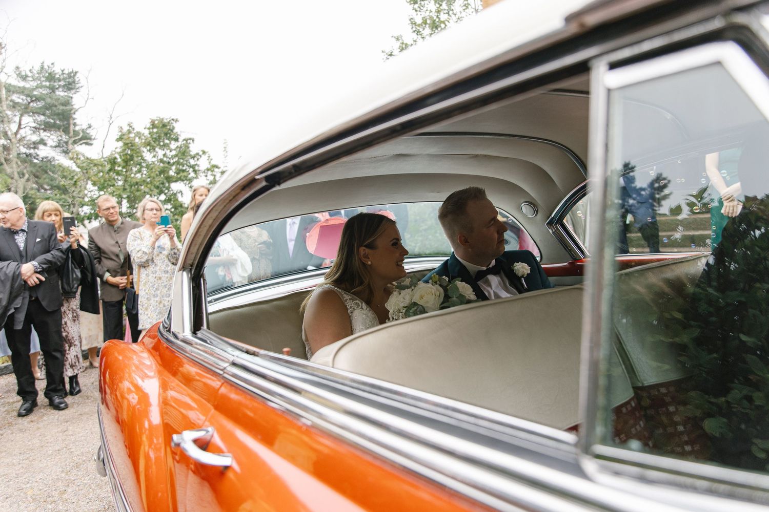 Wedding guests gathering around a classic orange car decorated for the celebration.