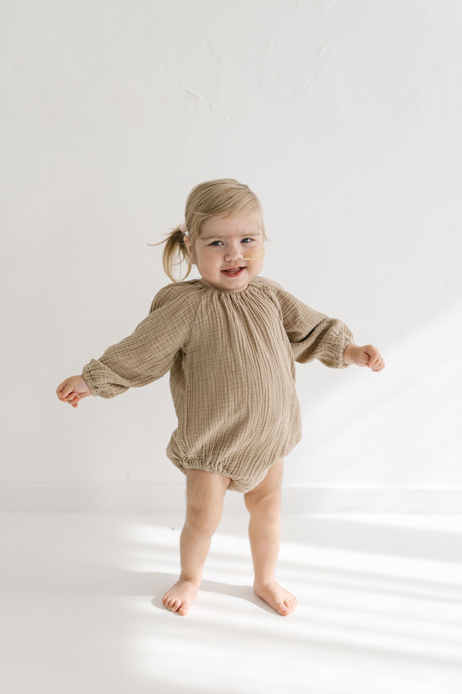 Baby in tan knit romper standing and playing against white background.