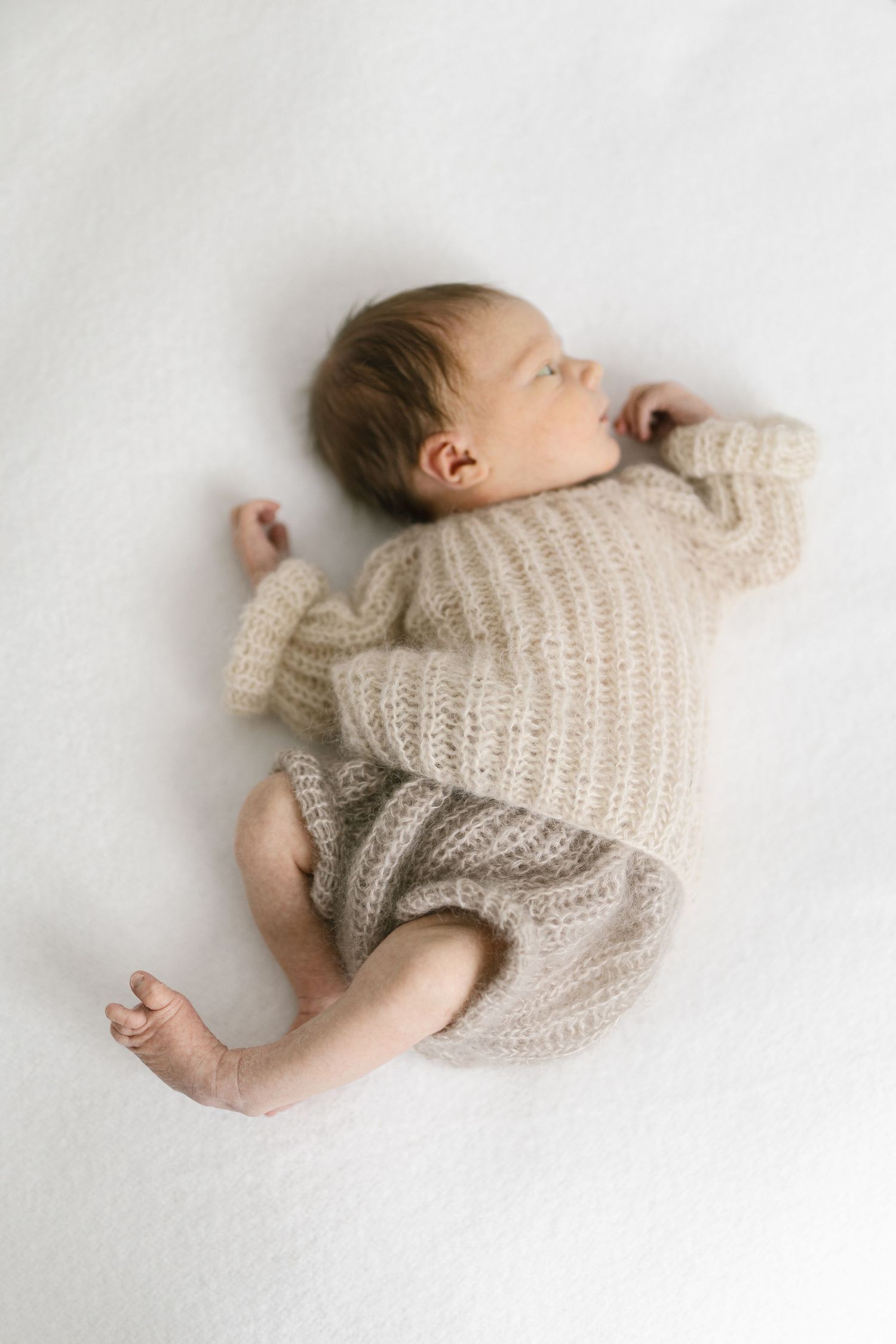 A sleeping newborn baby dressed in a cozy beige knit romper lies peacefully on a soft white blanket.