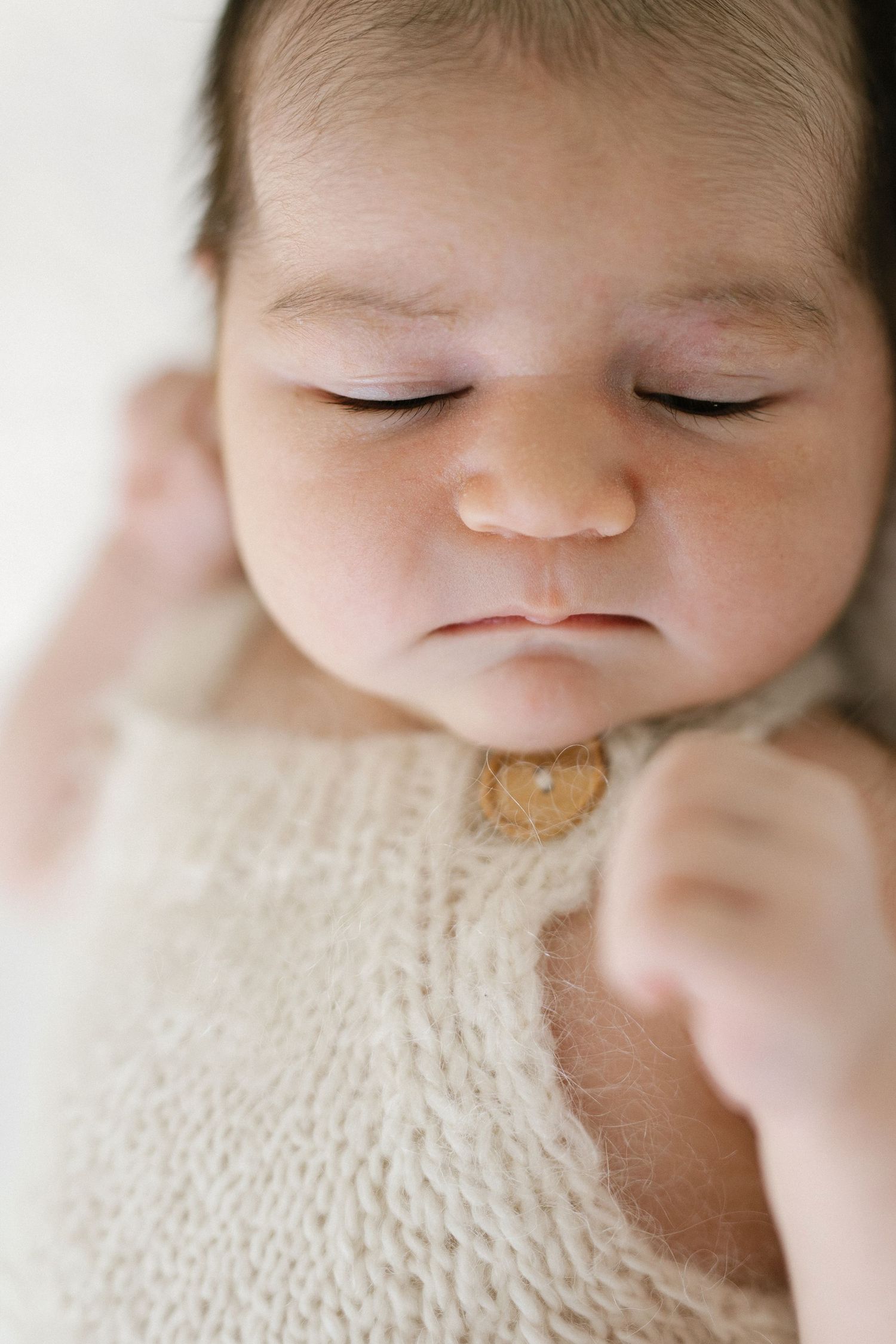 Sleeping newborn baby with a calm, pouty expression wearing a cream knit romper with a wooden button, tiny fists resting near the chest.