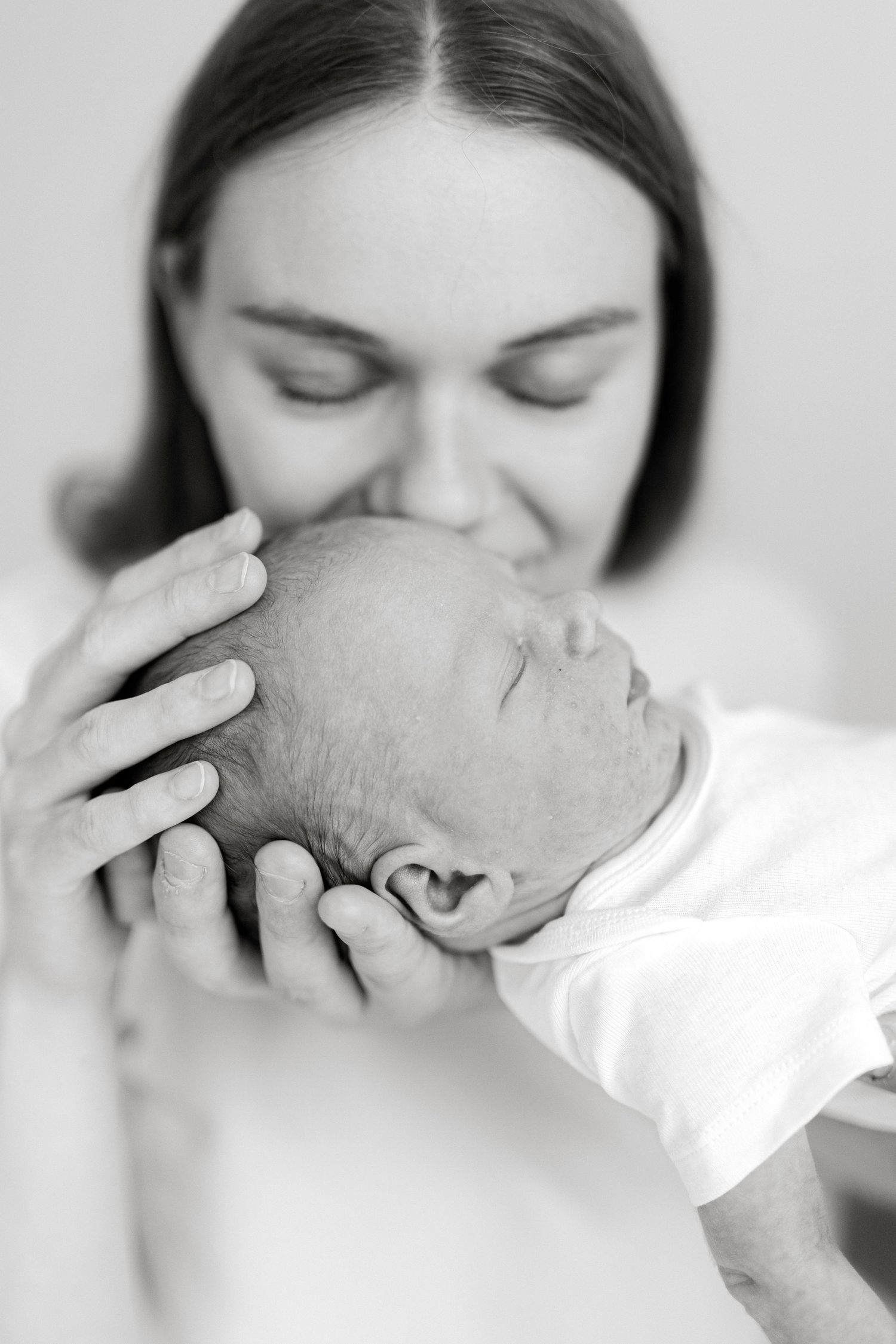 Black and white photo of a mother tenderly kissing her sleeping newborn baby's head, cradling it gently in her hands.
