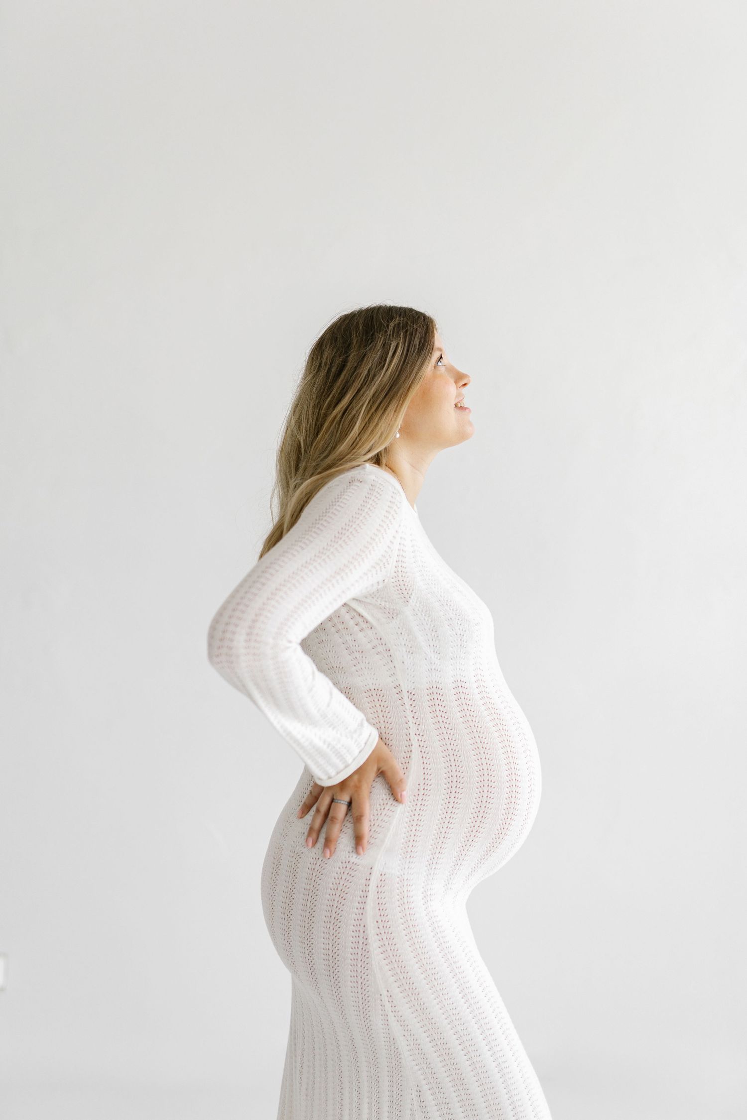 Pregnant woman in white lace dress posing sideways, hands on hips, against a white background.