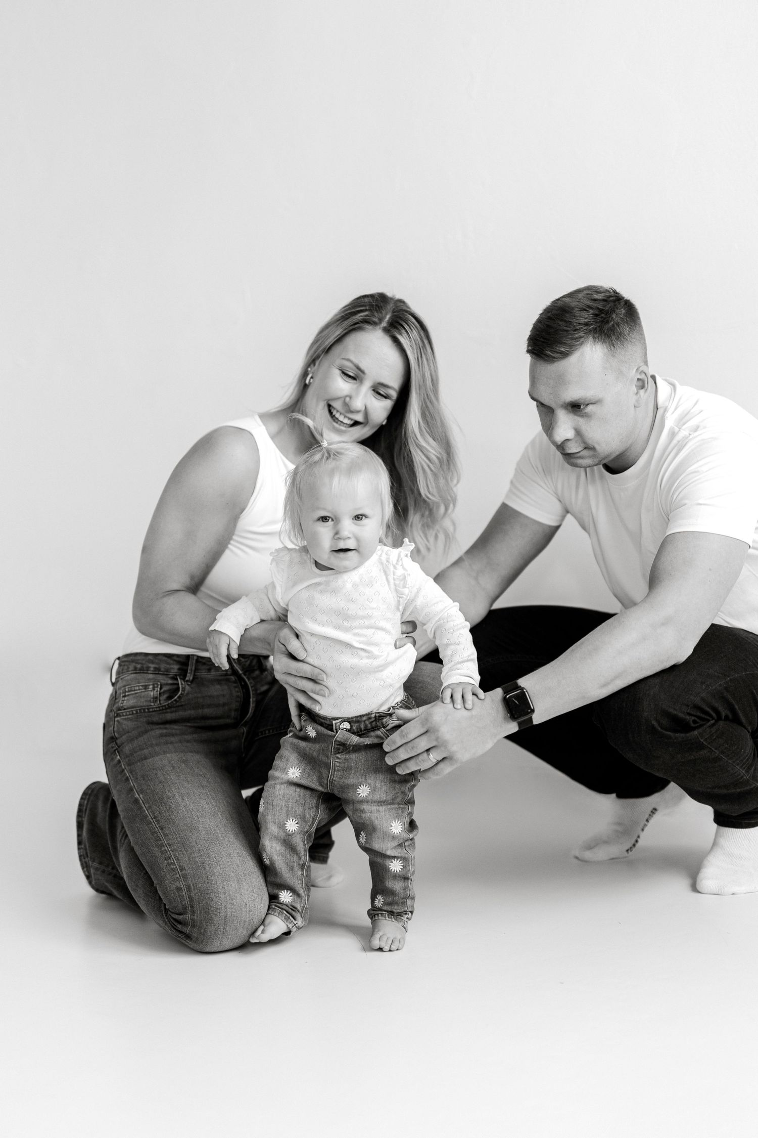 Black and white family photo of parents helping their baby take first steps in a studio setting.