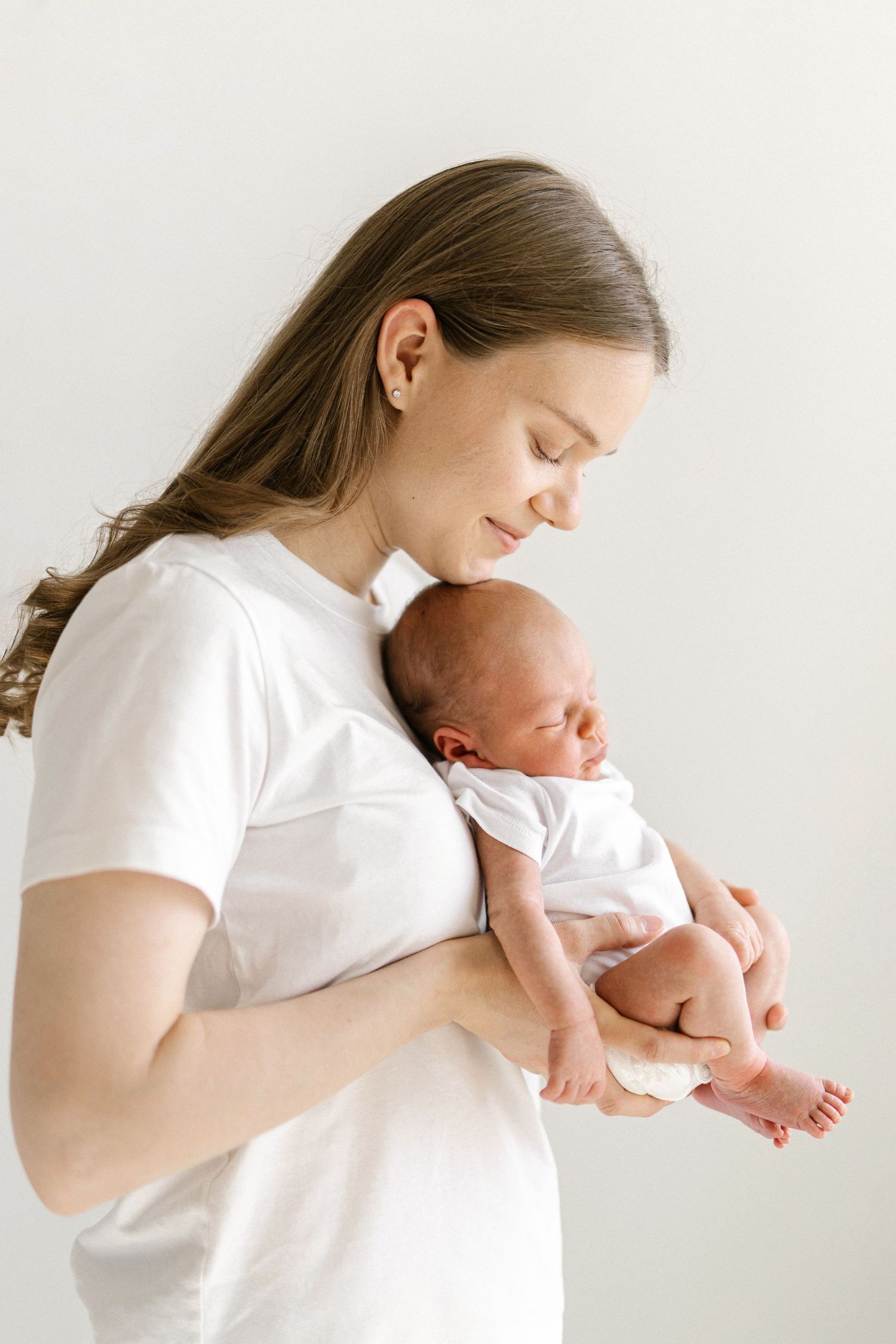 A young mother in a white t-shirt tenderly holds her sleeping newborn baby against a light background.