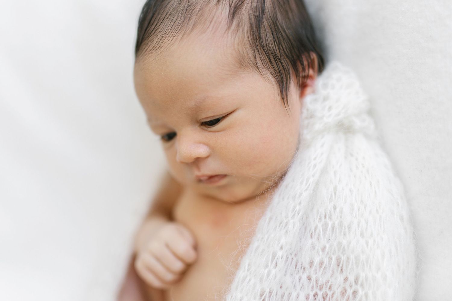 Peaceful newborn baby wrapped in white knit blanket, eyes closed, delicate features in soft natural light.