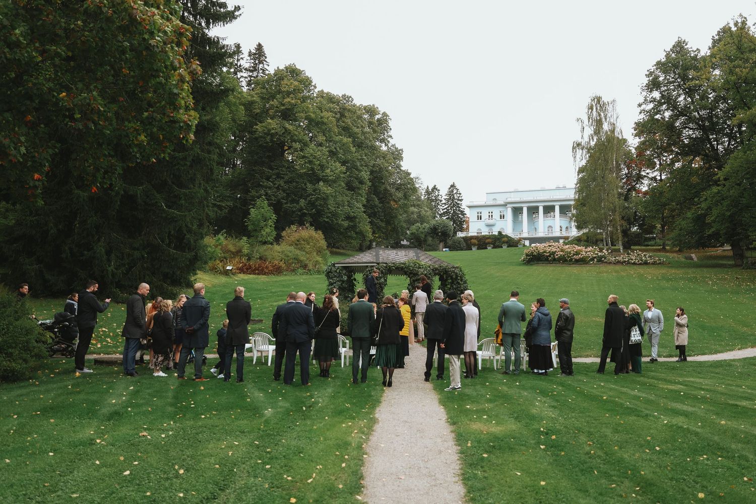 Guests gather on a green lawn near a white mansion surrounded by lush trees during an outdoor event.