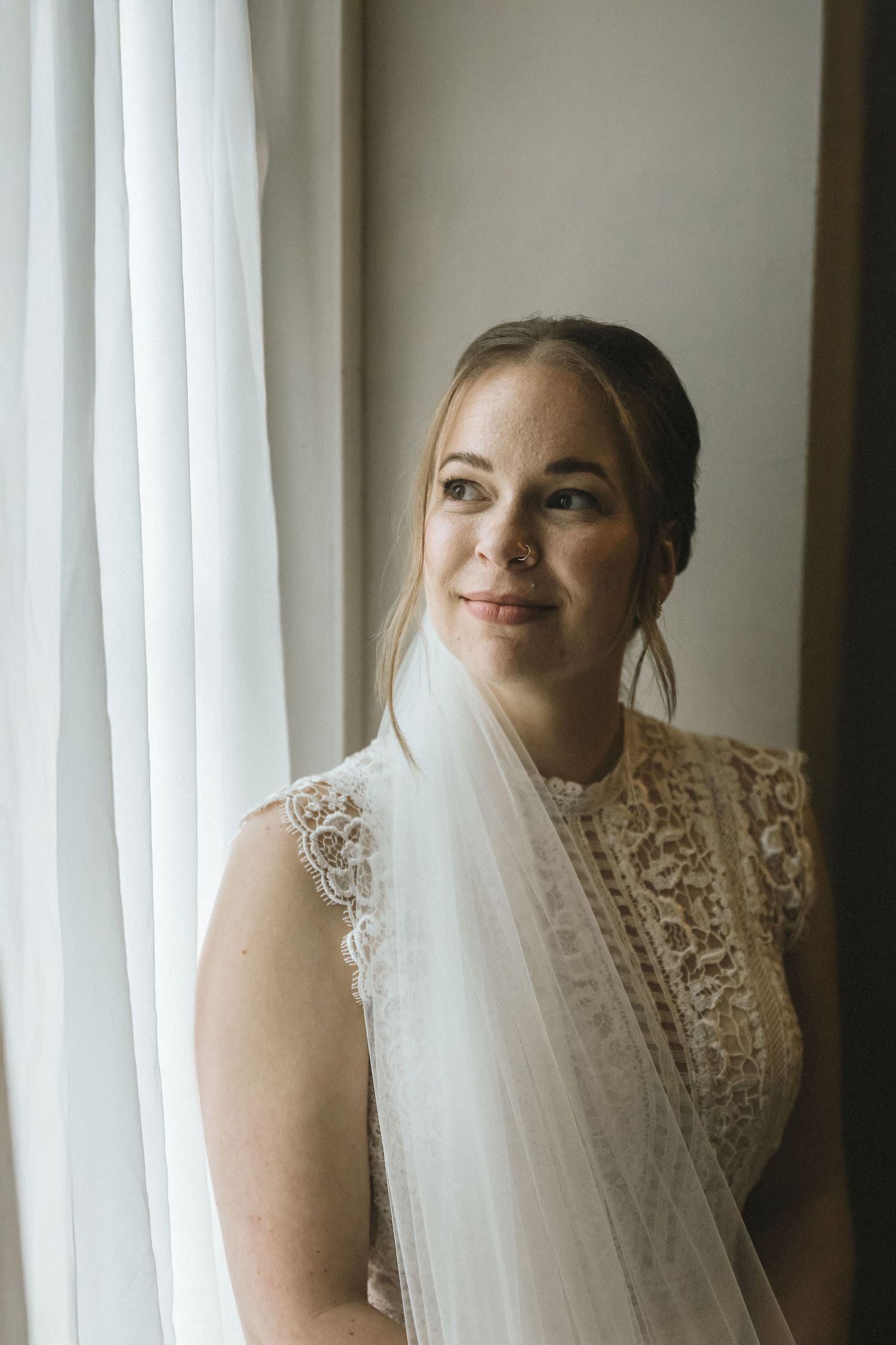 Elegant bride in lace wedding dress and veil stands by a window in soft natural light.