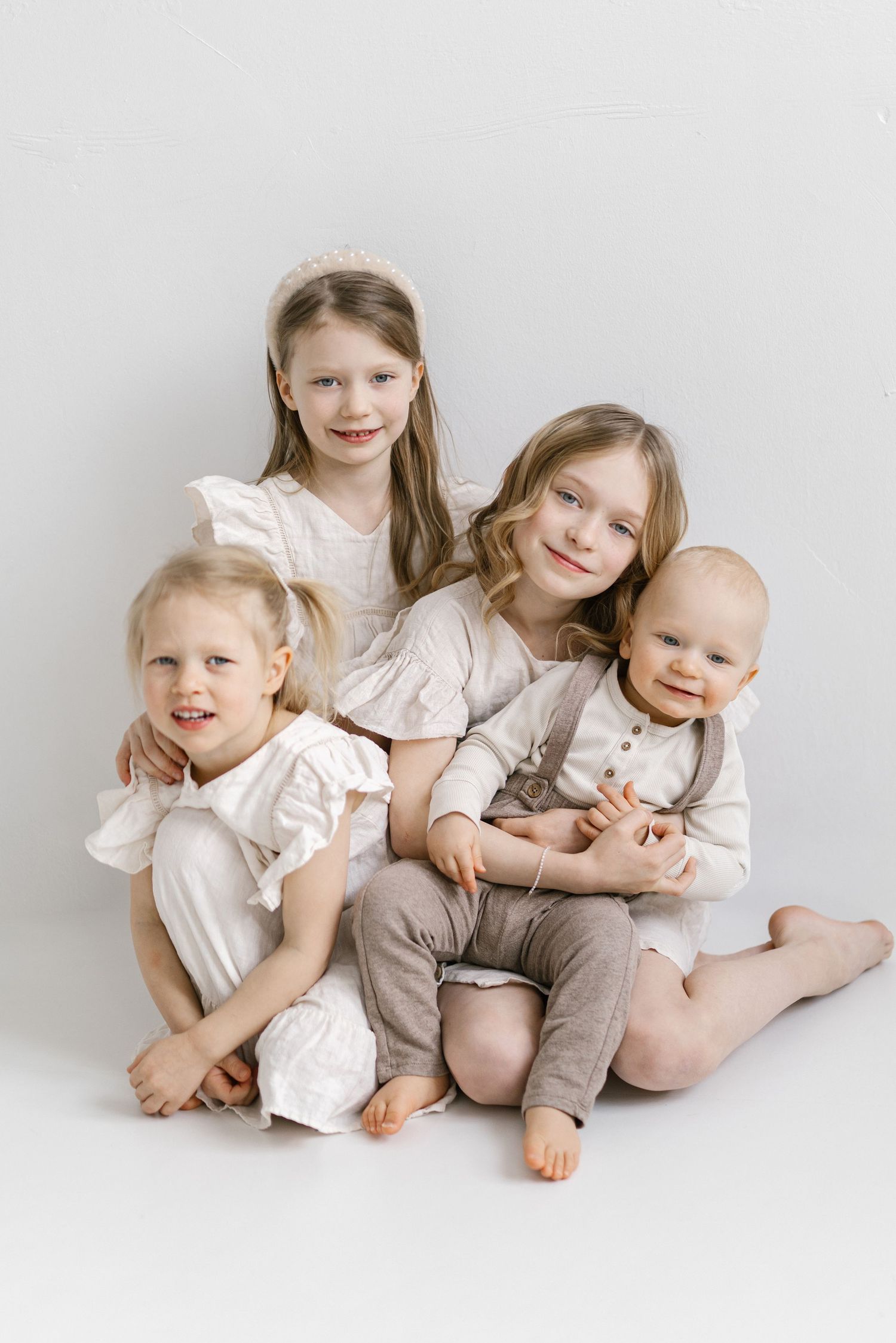 Four siblings wearing coordinated white and beige outfits sit together on a light background for a portrait.