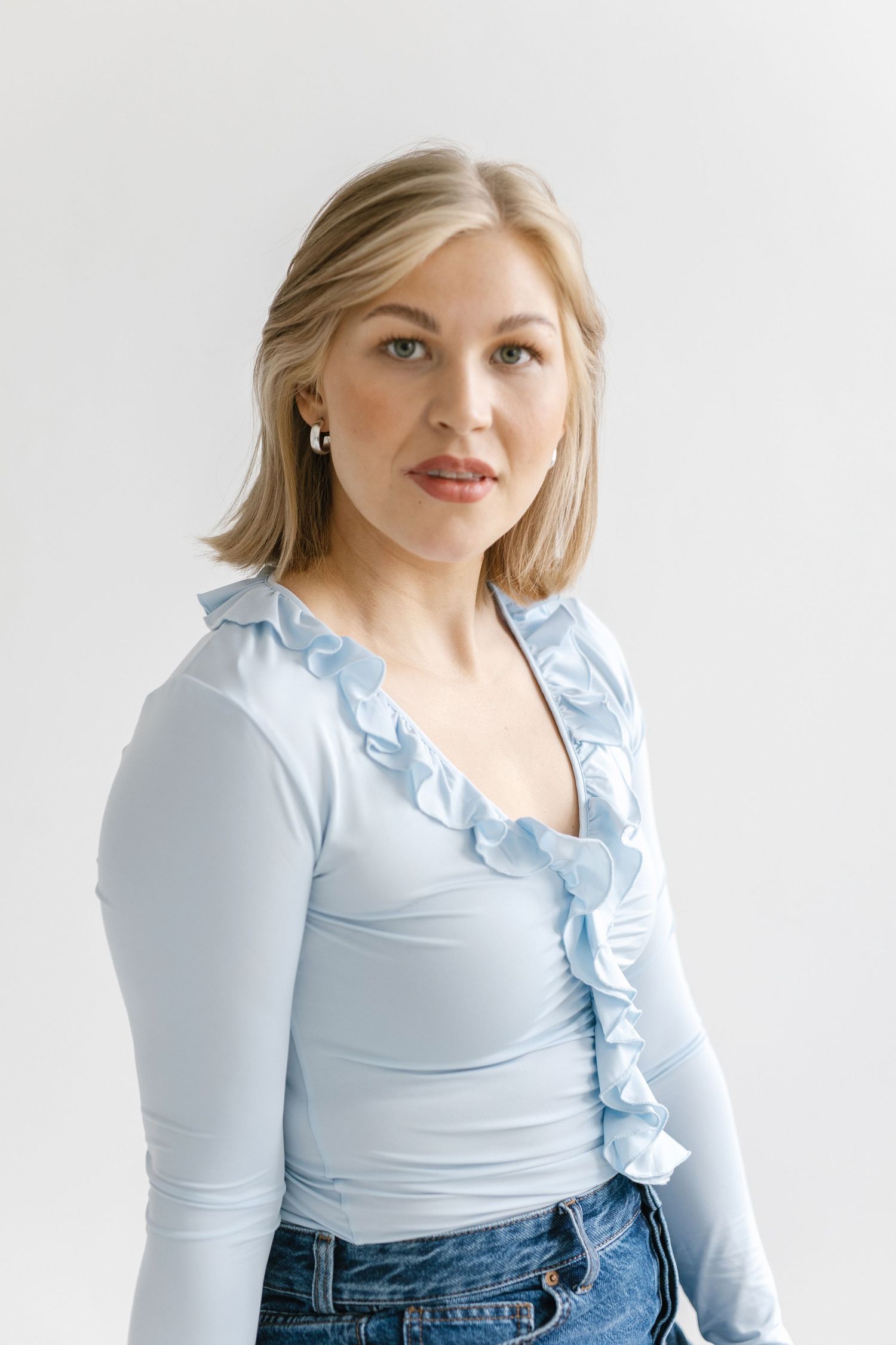A person wearing a light blue ruffled wrap top with blue jeans poses against a white background.
