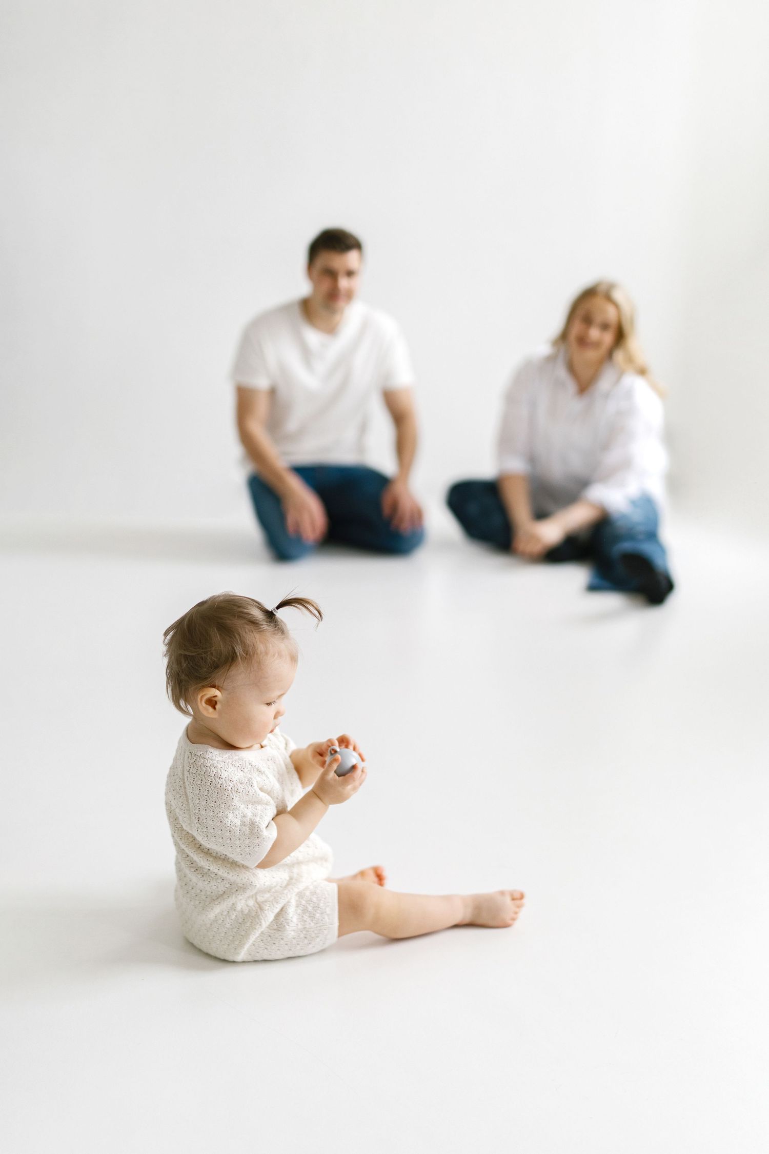 Happy family with baby girl sitting on white floor, parents watching toddler play in bright studio setting.