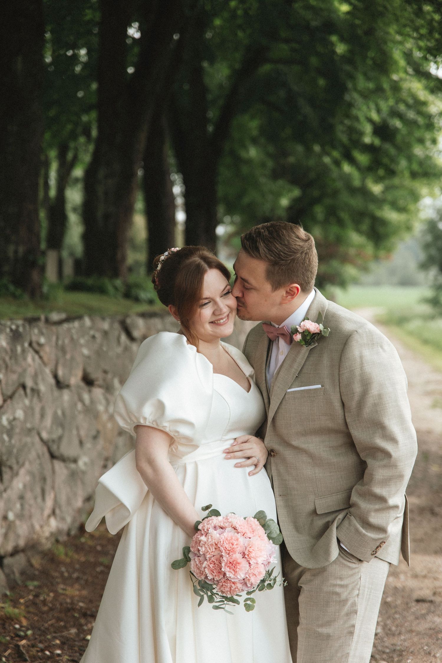 A groom in a beige suit kisses his bride on the cheek as she holds a pink bouquet on their wedding day.