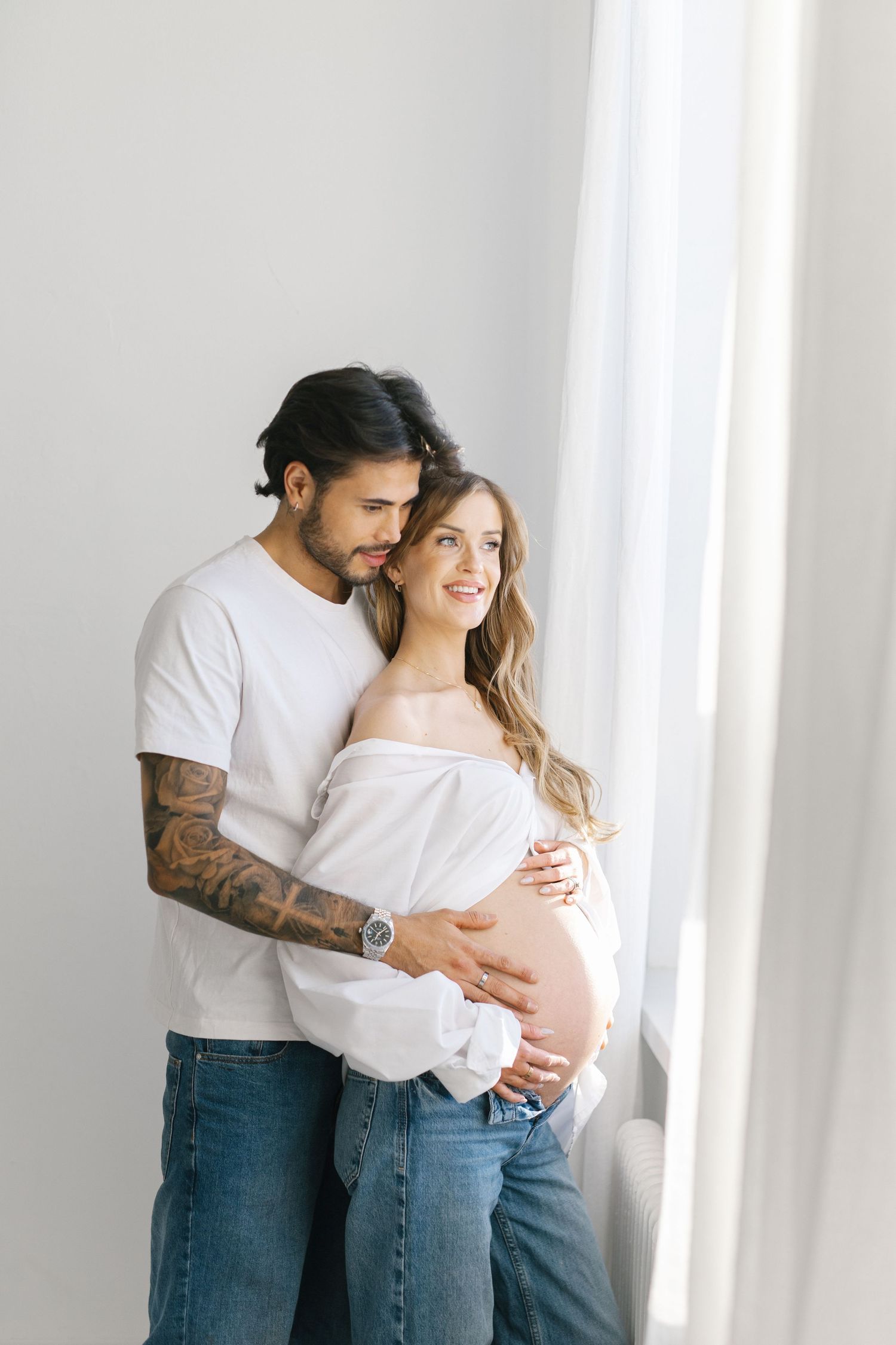 Couple embracing near window, pregnant woman in white off-shoulder top and jeans, tattooed man in white t-shirt.