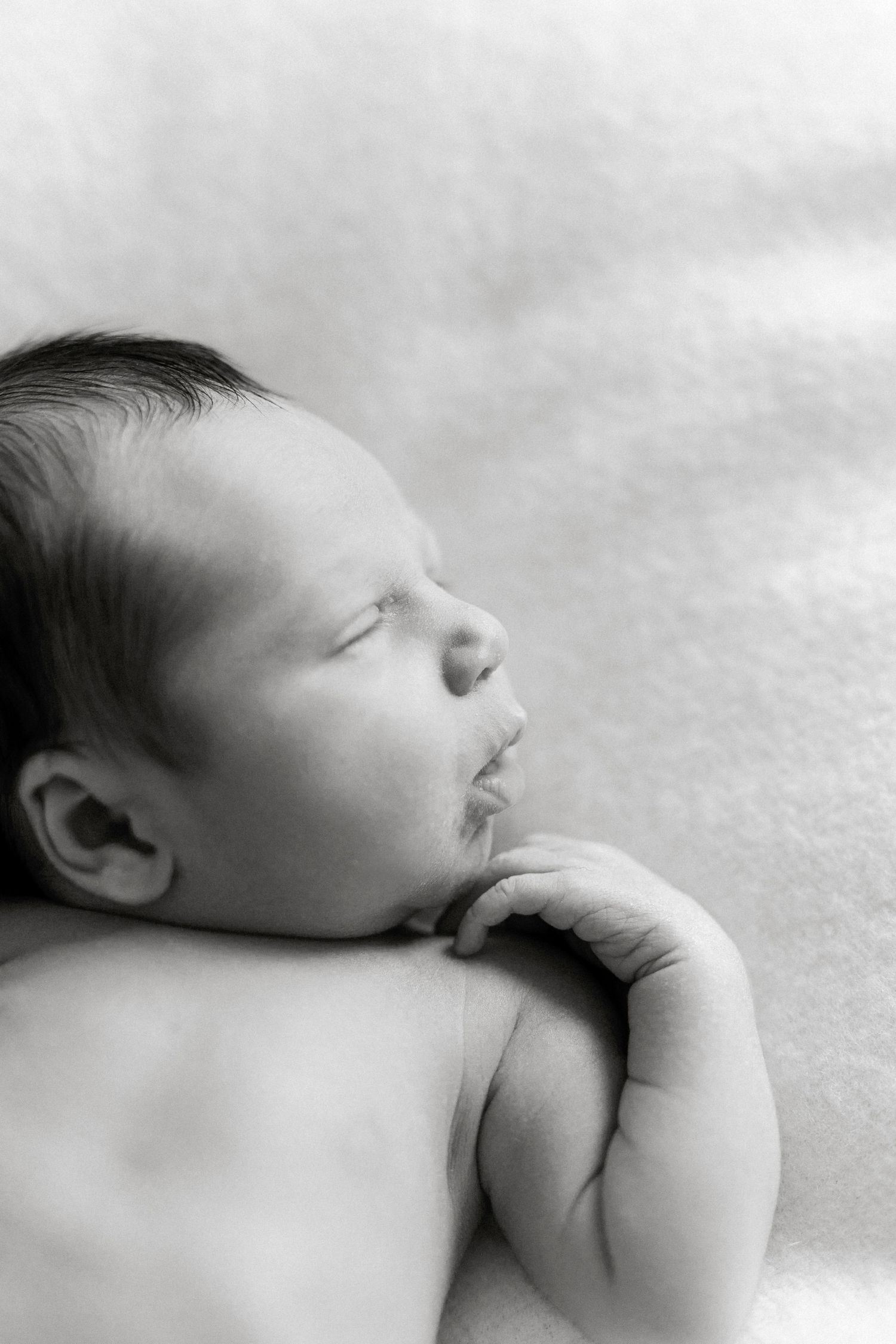 Black and white portrait of a peaceful sleeping newborn baby with dark hair, hands tucked under chin.