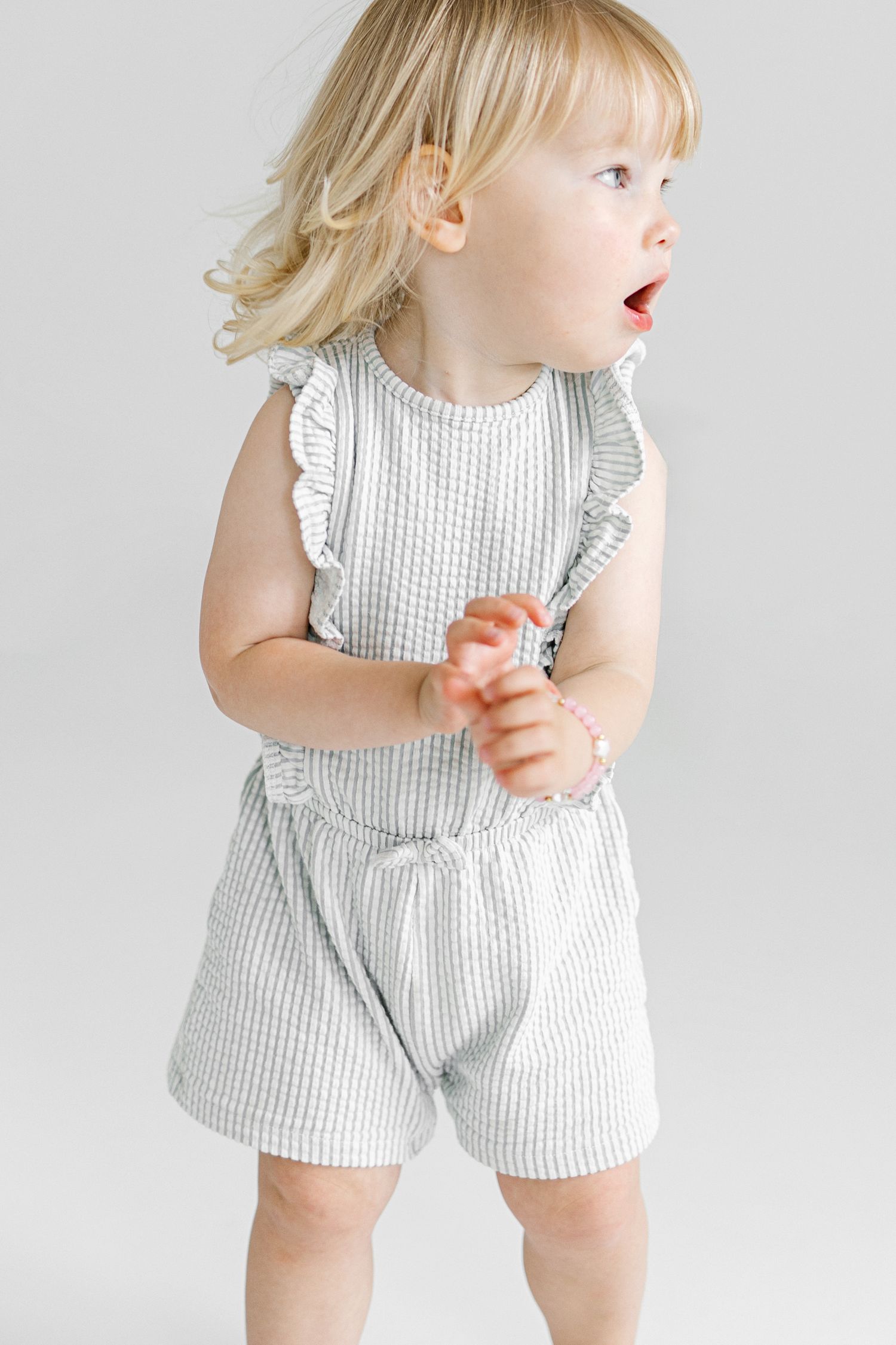 Toddler girl in a gray striped ruffle romper with shorts, blonde hair, mouth open in surprise.