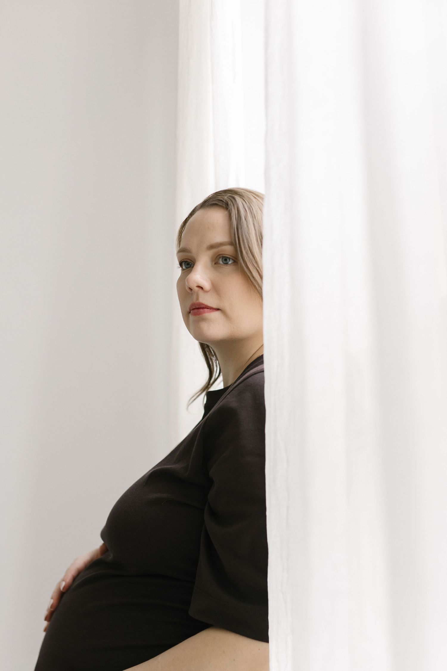 Pregnant woman in black dress holding baby bump near white curtains.