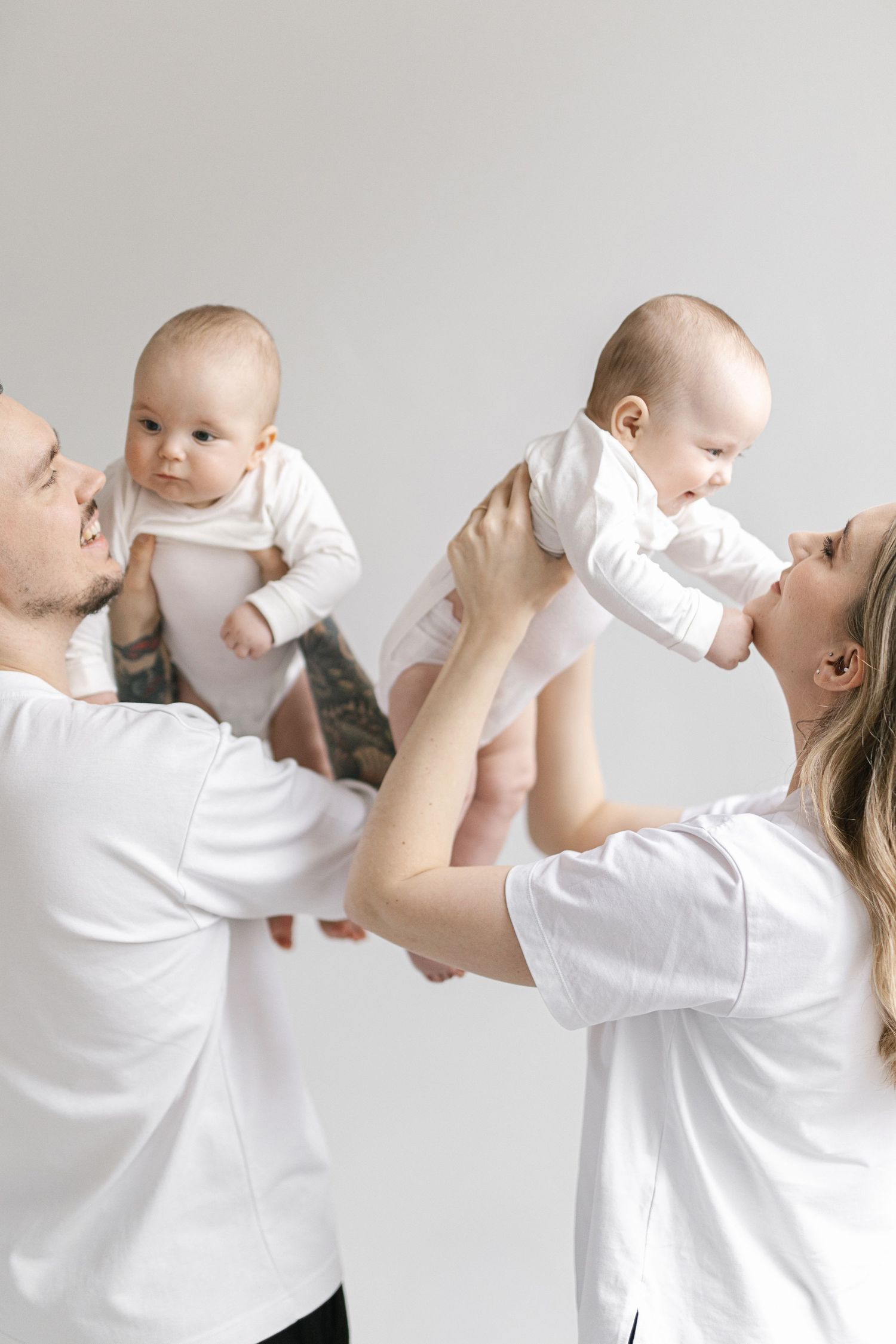 Parents in white shirts joyfully lift twin babies dressed in white onesies against a light gray background.