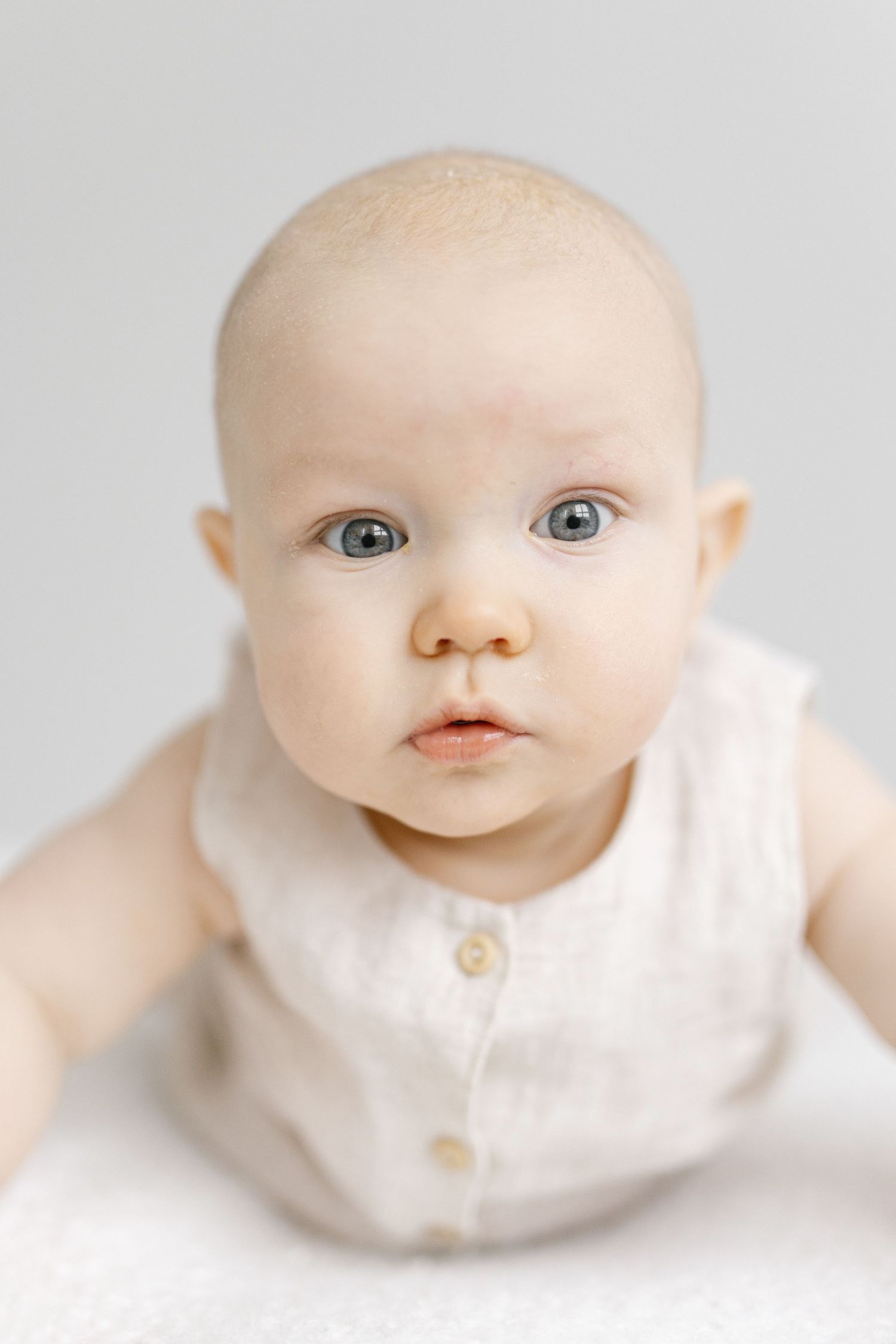 Adorable baby with blue eyes wearing a white linen outfit, looking curiously at the camera against a light background.