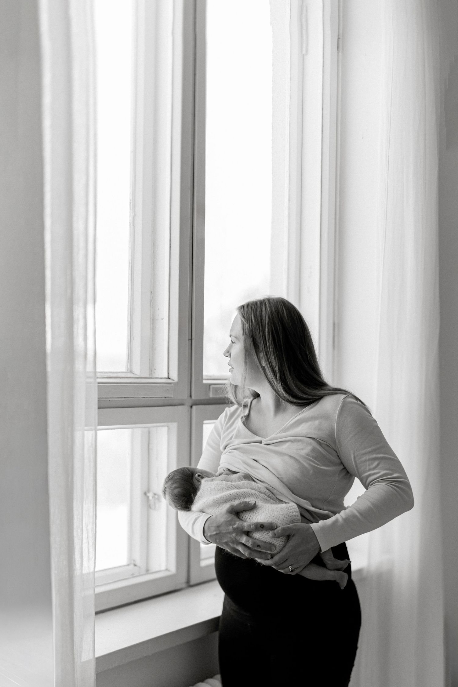 Black and white photo of a mother holding her newborn baby while standing by a bright window.