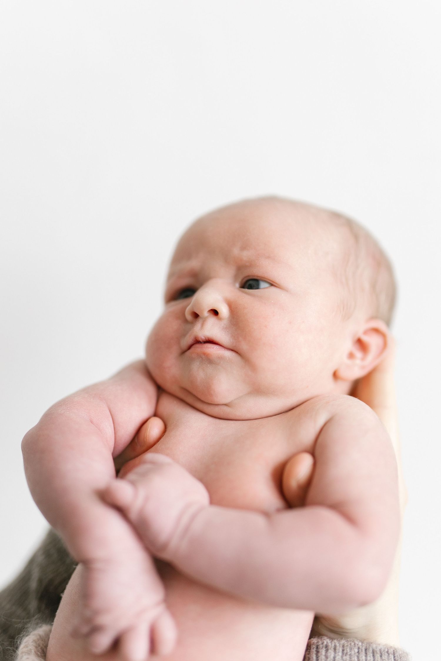 A newborn baby with chubby cheeks and folded arms gazes upward against a clean white background.