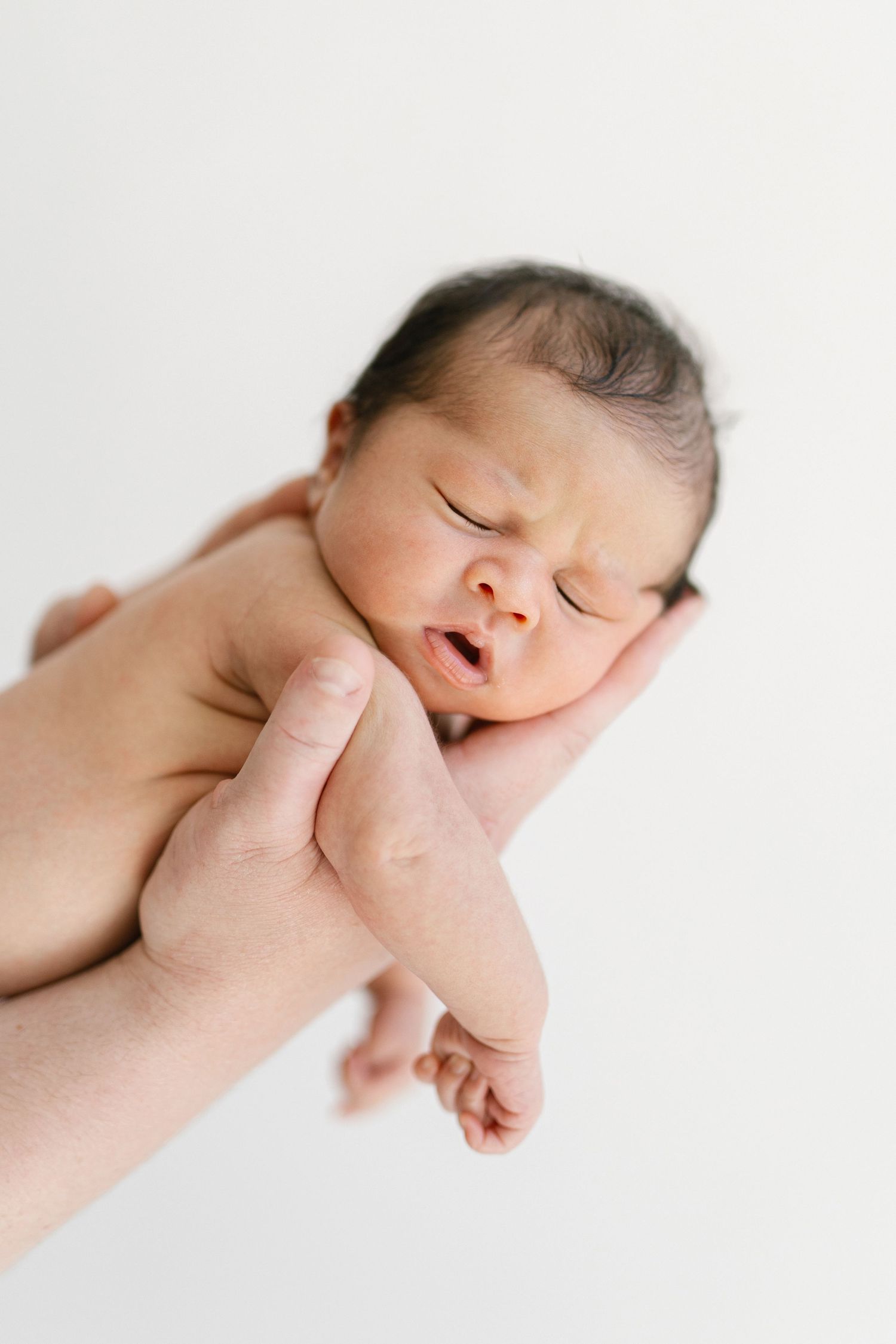 Newborn baby with dark hair sleeping peacefully while being cradled in an adult's hand against white background.