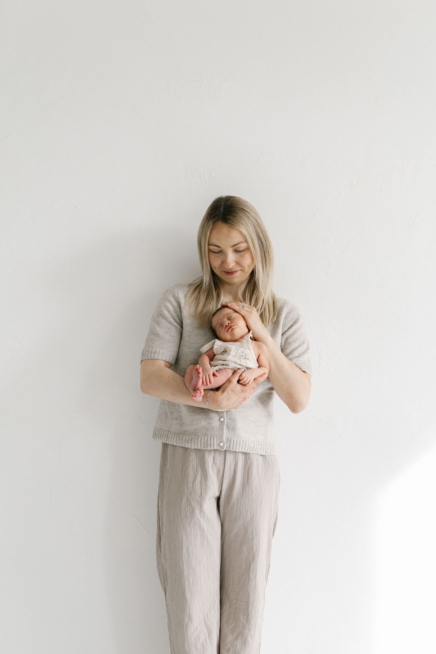A young blonde woman in neutral linen outfit gently holds a sleeping newborn baby against a white wall.