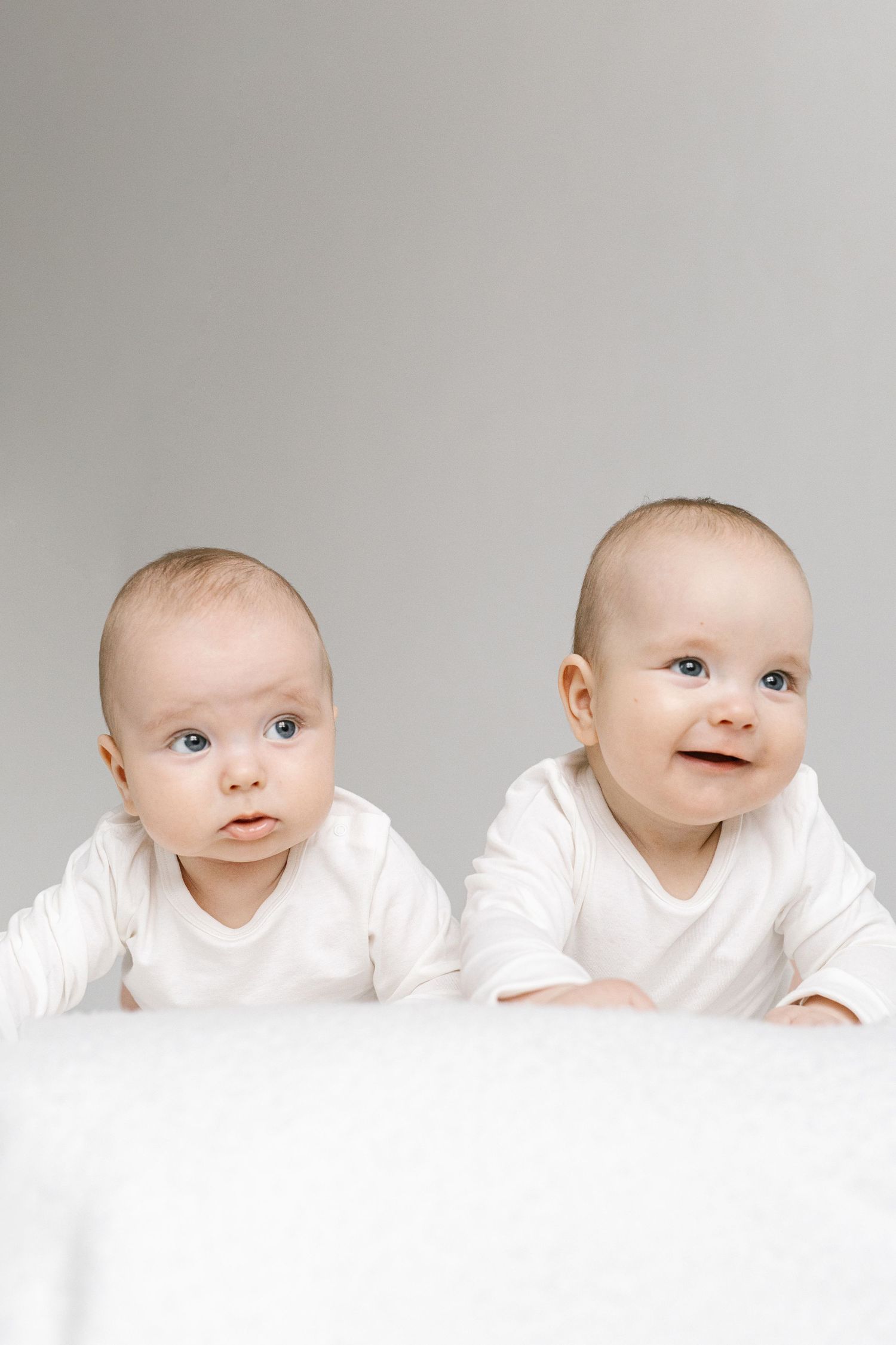 Two adorable blue-eyed twin babies in white onesies lying on a white surface against a gray background.