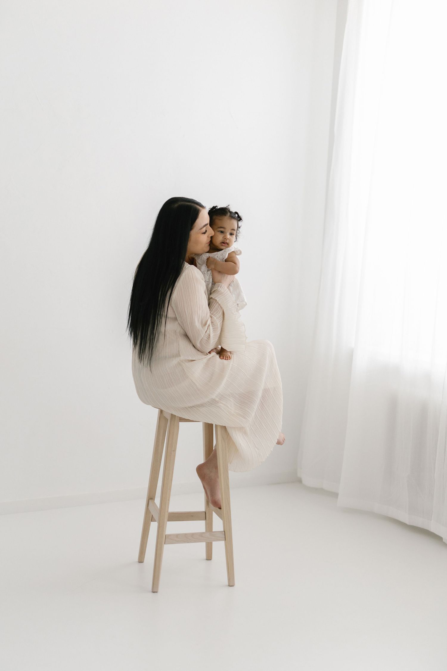 A mother in a beige dress sits on a wooden stool kissing her baby in a bright white studio.