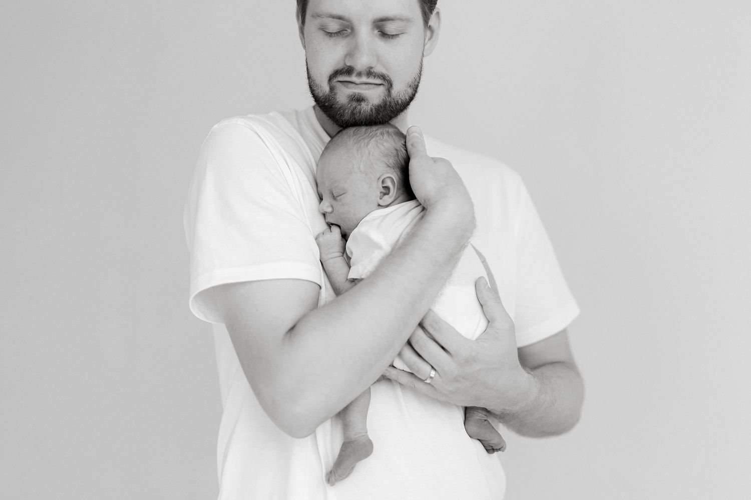 Black and white photo of a bearded father tenderly holding his sleeping newborn baby against his chest.