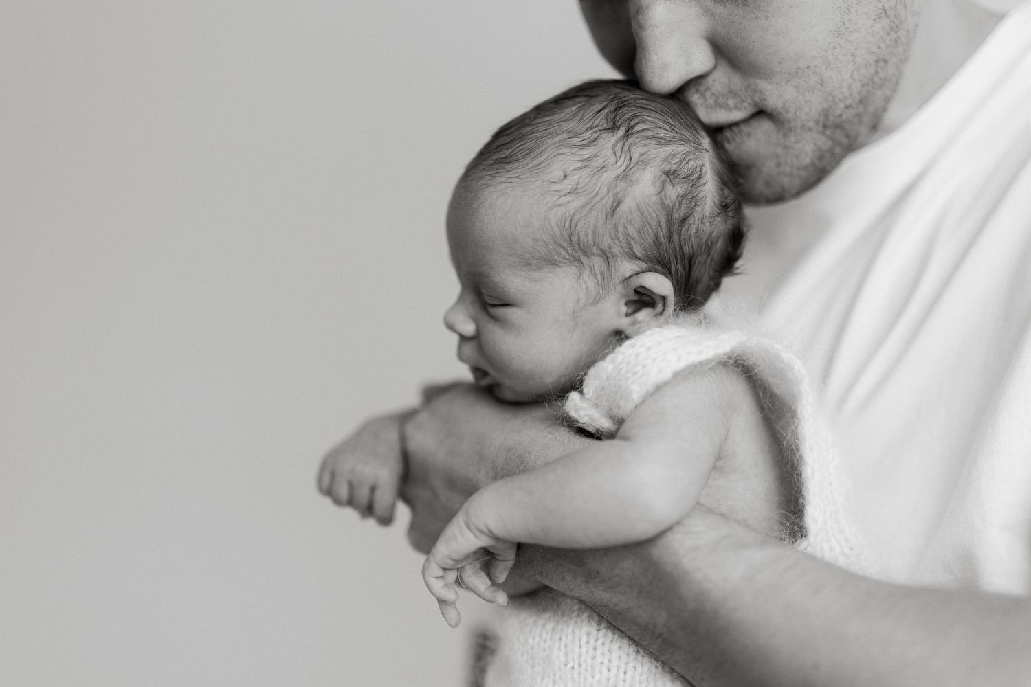 Black and white photo of a father tenderly kissing his newborn baby while cradling the infant in his arms.
