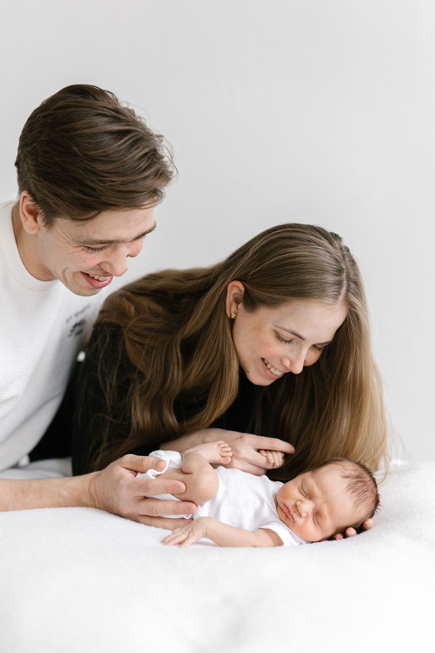Young parents smiling lovingly at their sleeping newborn baby lying on a white bed.