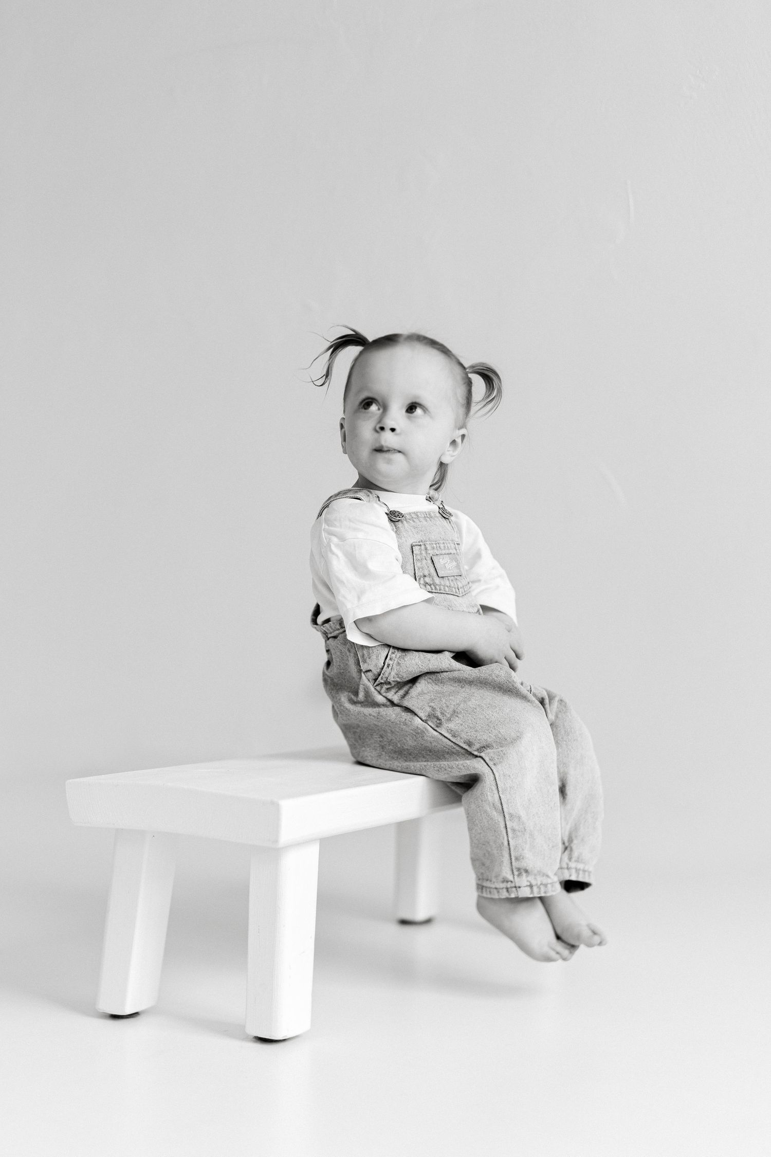 Black and white photo of a cute toddler girl in denim overalls sitting on a small white bench with arms crossed.
